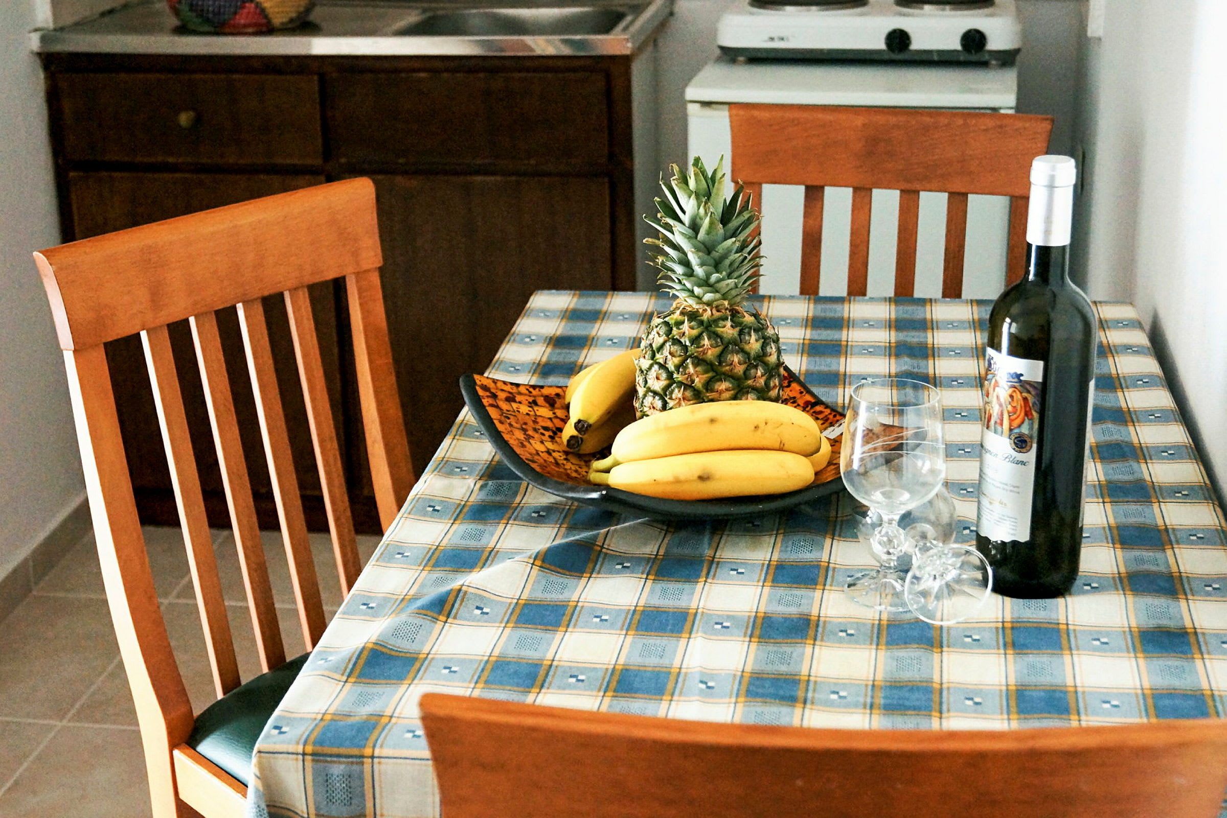 Keittiön pöytä, jolla on hedelmäkulho, jossa on ananas ja banaaneja, viinipullo ja kaksi viinilasia. (Tekoälyn luoma teksti)