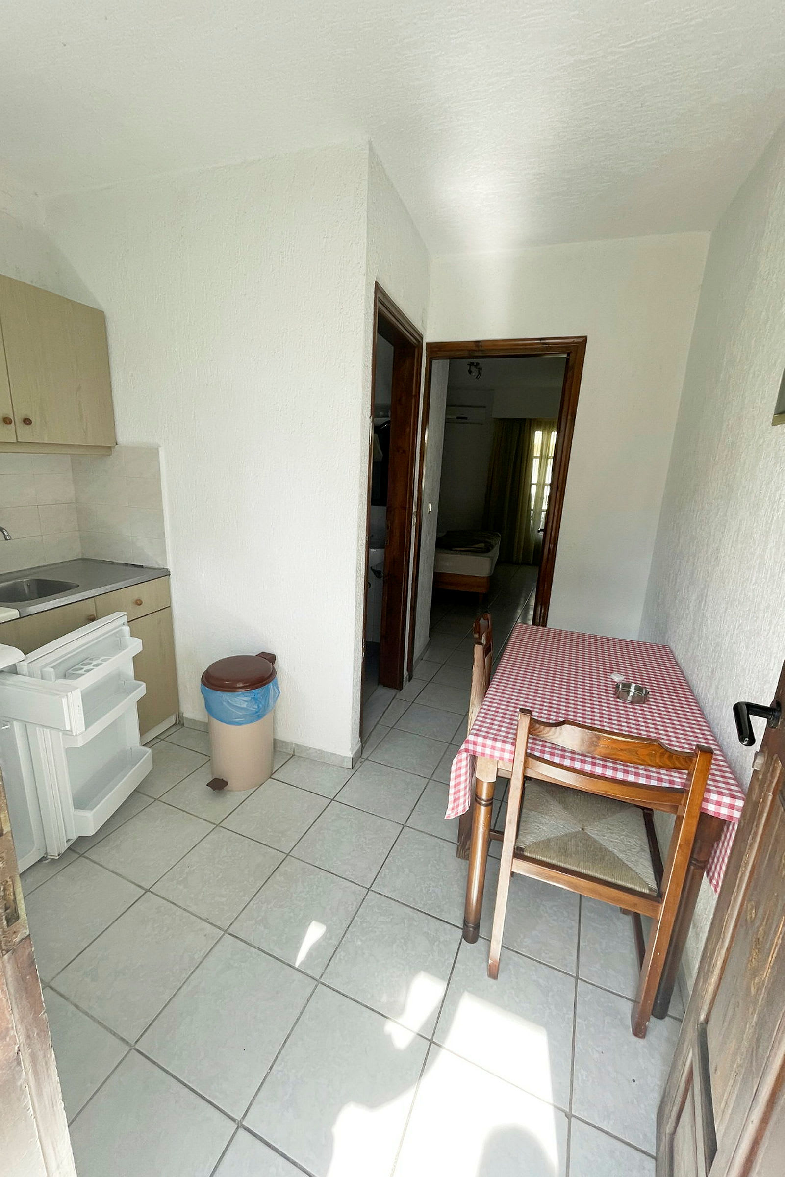 Keittiö, jossa on punaruutuinen pöytäliina, yksi tuoli ja avoin jääkaappi. Ovi johtaa makuuhuoneeseen. (Tekoälyn luoma teksti)