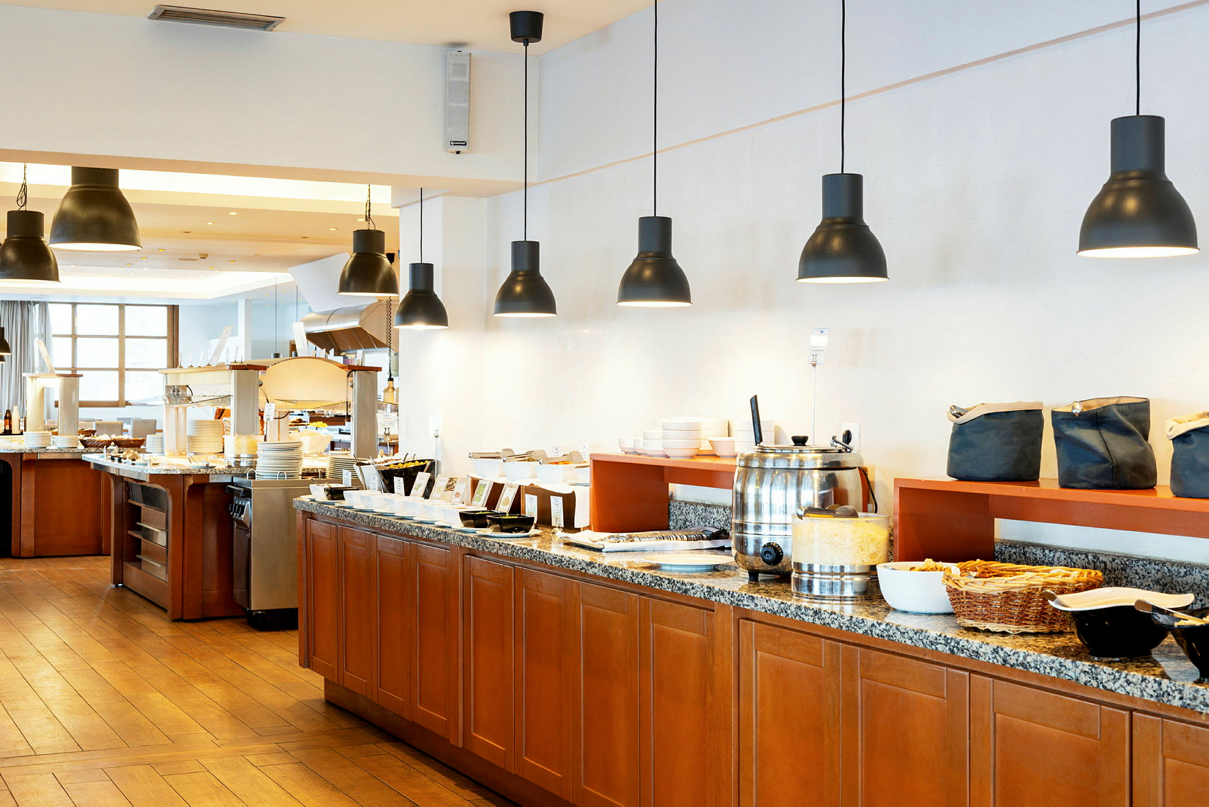 Buffet-alue, jossa ruokia ja juomia on aseteltu pitkälle tiskille, jossa on puukaapit, ja jota valaisee useita mustia kattovalaisimia. (Tekoälyn luoma teksti)