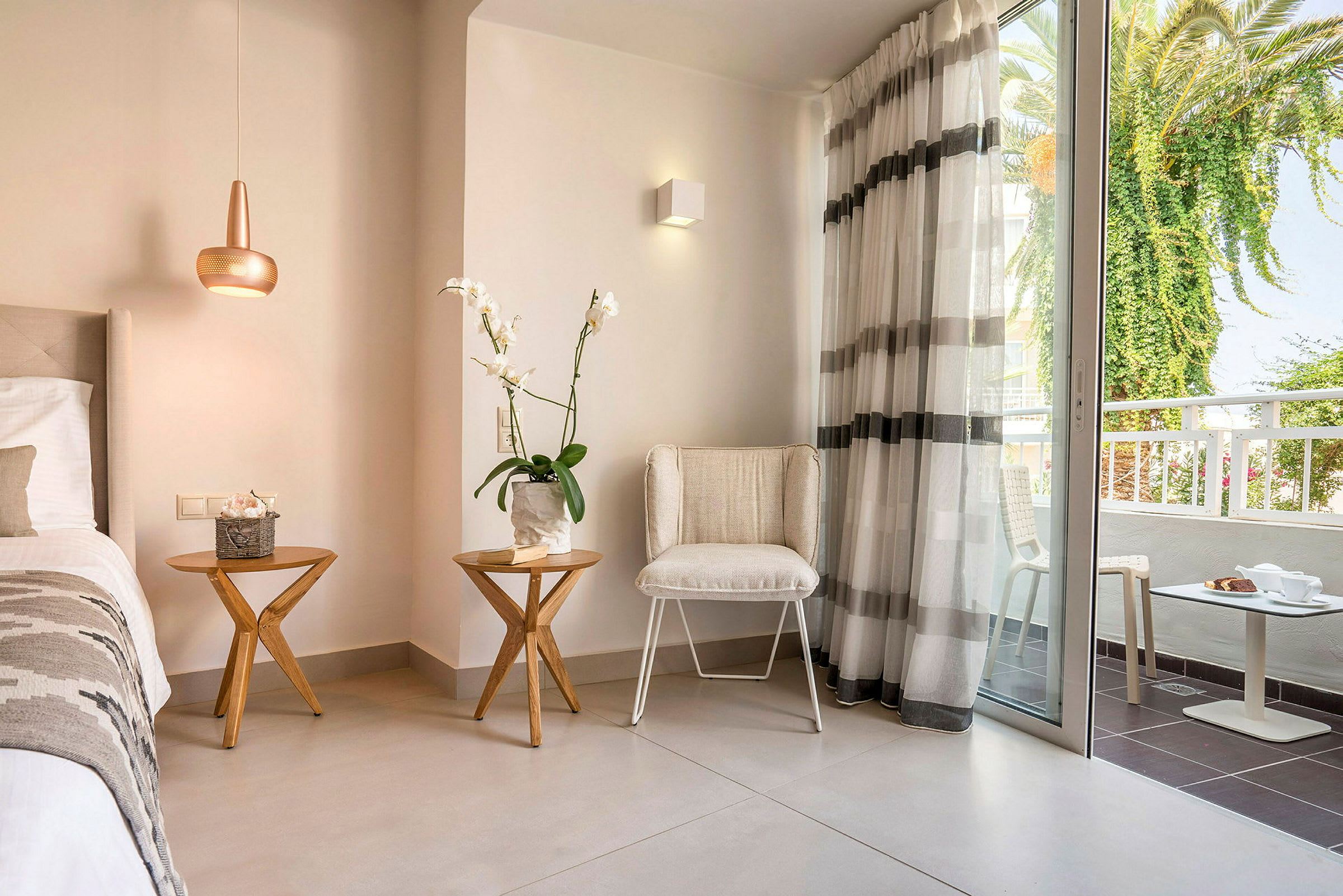 Hotellihuone, jossa on kaksi pöytää, tuoli, sänky ja lasiovi, joka johtaa parvekkeelle. (Tekoälyn luoma teksti)