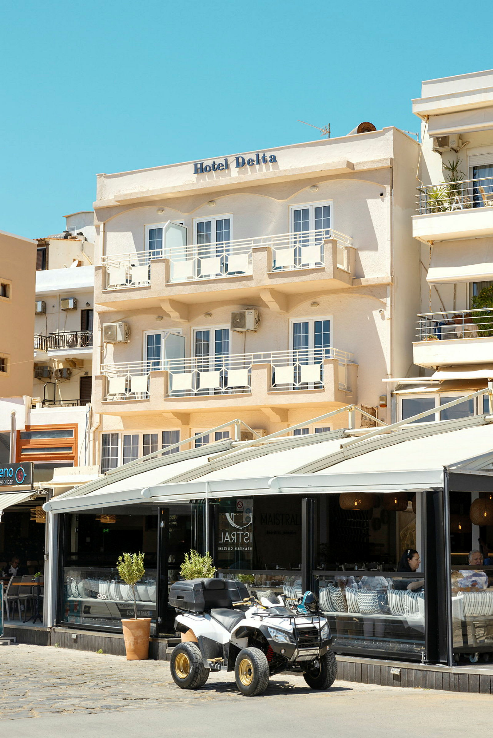 Hotellin julkisivu nimeltä "Hotel Delta", jossa on parvekkeita ja kahvila pohjakerroksessa, ulkopuolella on pysäköitynä nelipyöräinen ajoneuvo. (Tekoälyn luoma teksti)