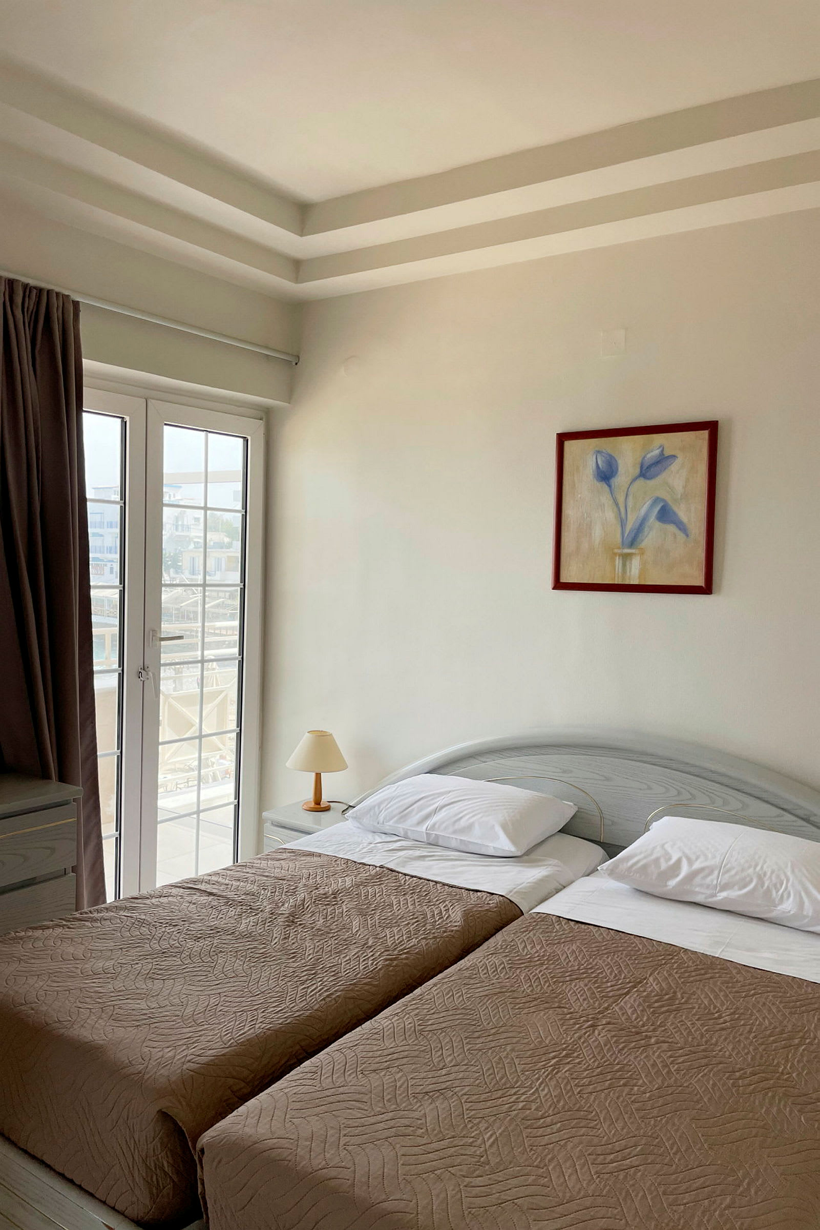 Hotellihuone, jossa on kaksi yhden hengen vuodetta, lamppu ja lasiovi. (Tekoälyn luoma teksti)