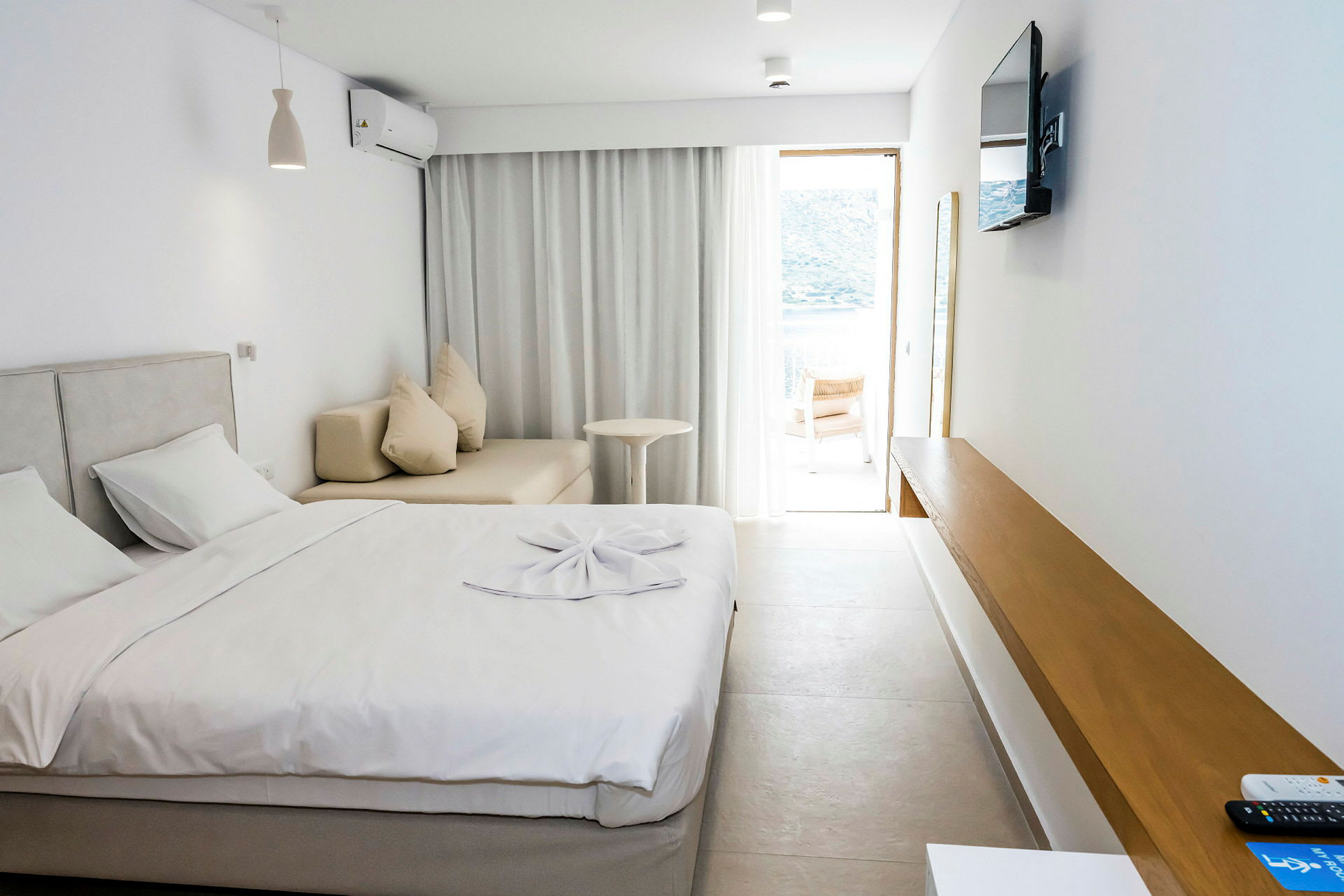 Hotellihuone, jossa on sänky, sohva, pöytä ja seinällä televisio. Ikkuna vaaleine verhoineen avautuu terassille. (Tekoälyn luoma teksti)