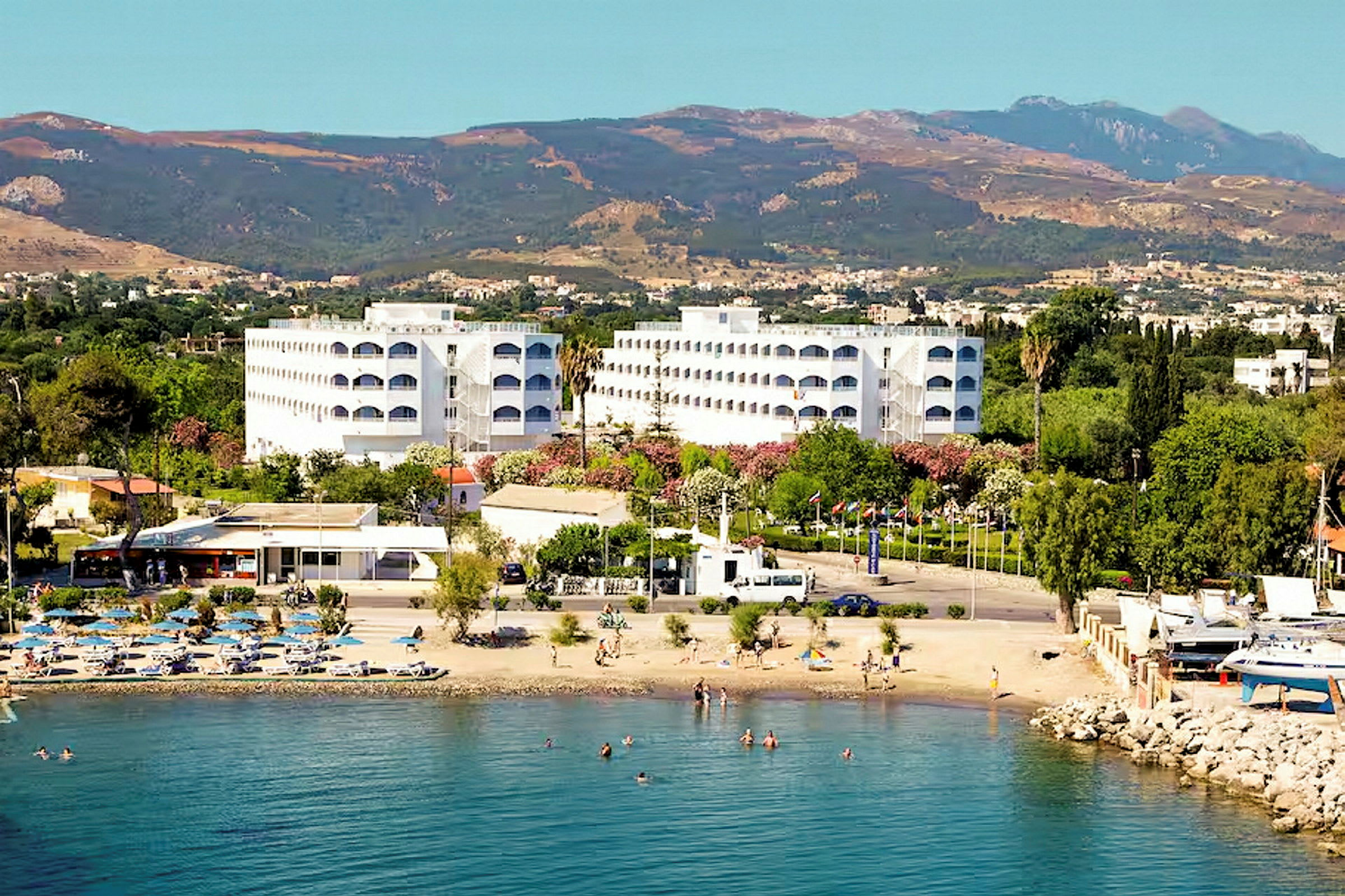 Hotellirakennuksia rannalla, uimareita ja vuoria taustalla. (Tekoälyn luoma teksti)