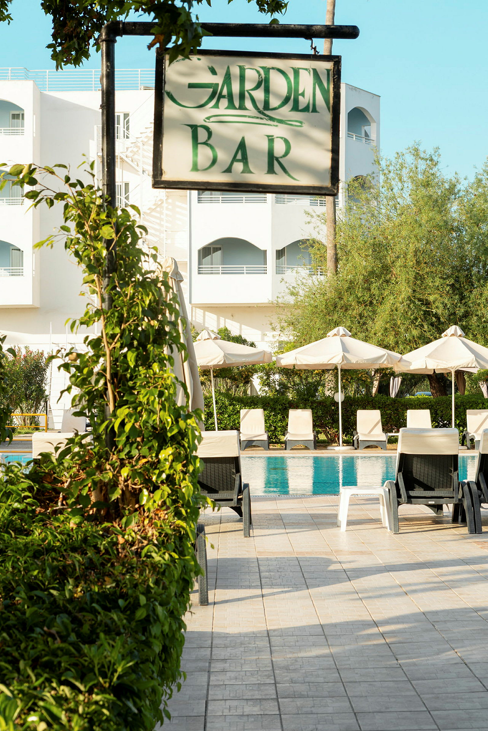 Kyltti, jossa lukee 'Garden Bar', uima-allasalueen ja hotellirakennuksen edessä. (Tekoälyn luoma teksti)
