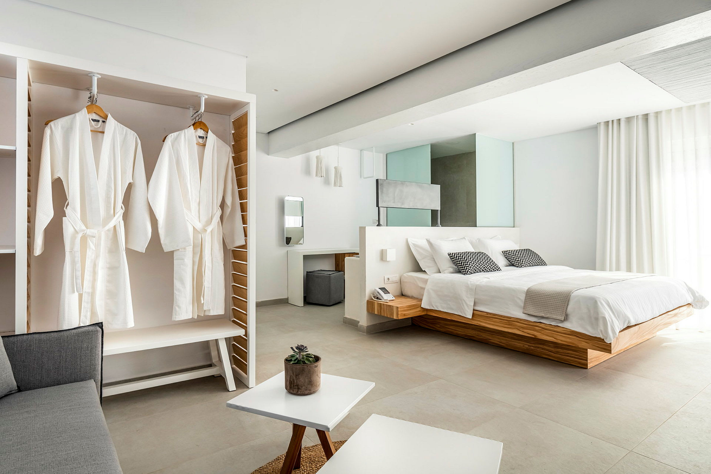 Kuva tyylikkäästä hotellihuoneesta, jossa on suuri sänky, vaatekaapissa roikkuvat valkoiset kylpytakit ja moderneja vaaleita huonekaluja. (Tekoälyn luoma teksti)