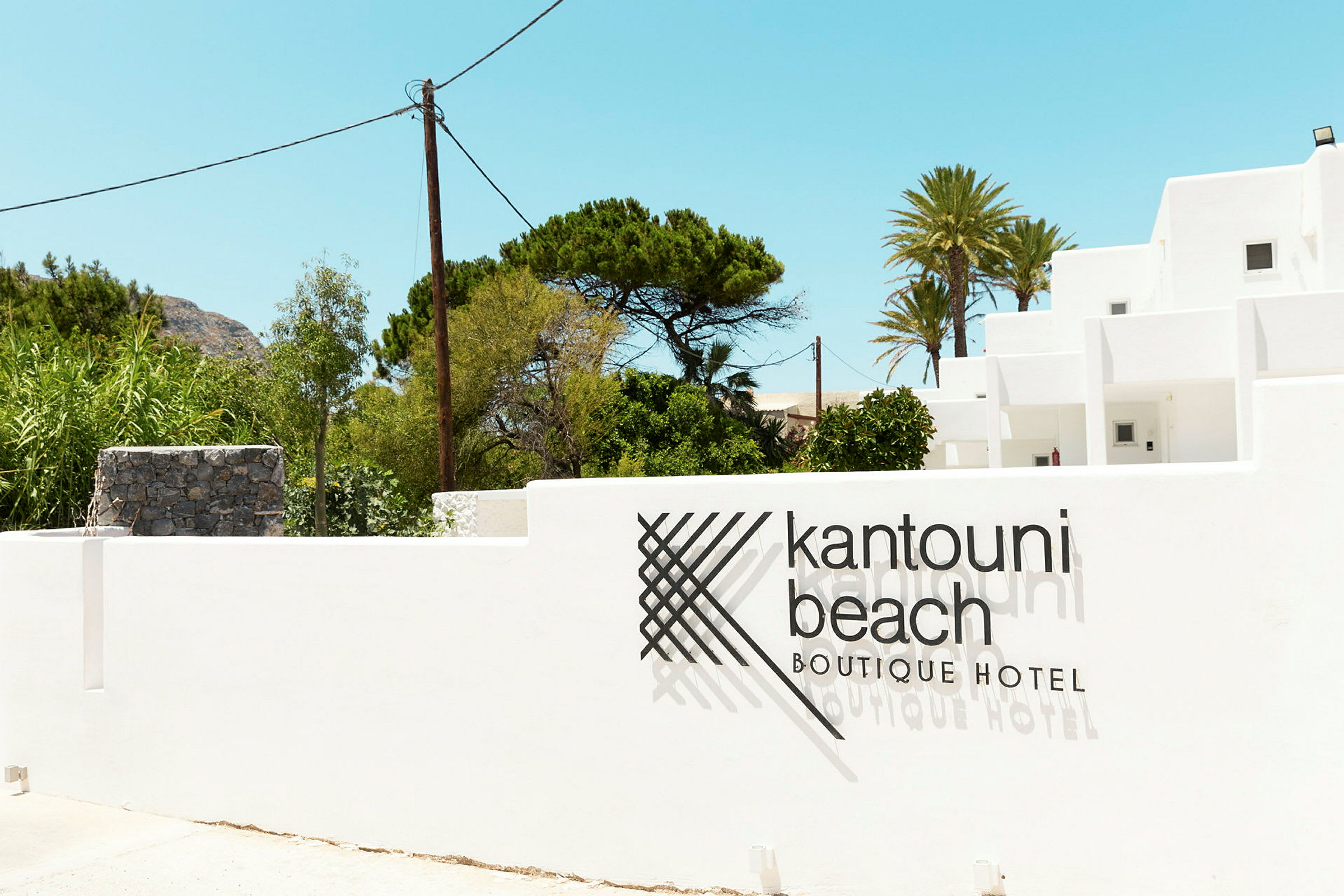 Kuvassa valkoinen rakennus, jonka edessä on muurissa teksti 'Kantouni Beach Boutique Hotel'. Taustalla vehreyttä ja kirkas taivas. (Tekoälyn luoma teksti)
