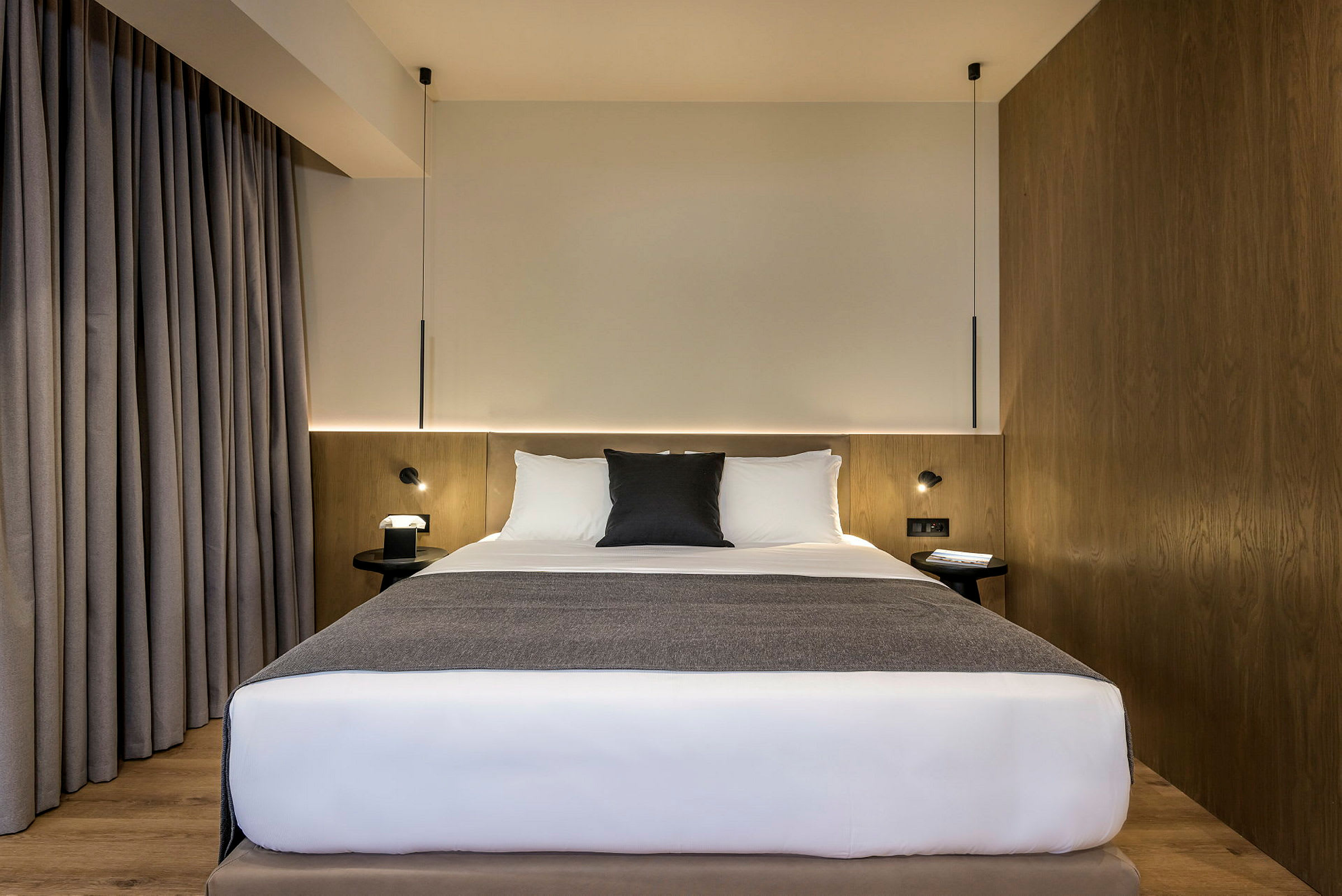 Hotellihuone, jossa on sänky valkoisilla lakanoilla ja harmailla yksityiskohdilla, puiset seinäpaneelit sekä harmaat verhot. (Tekoälyn luoma teksti)