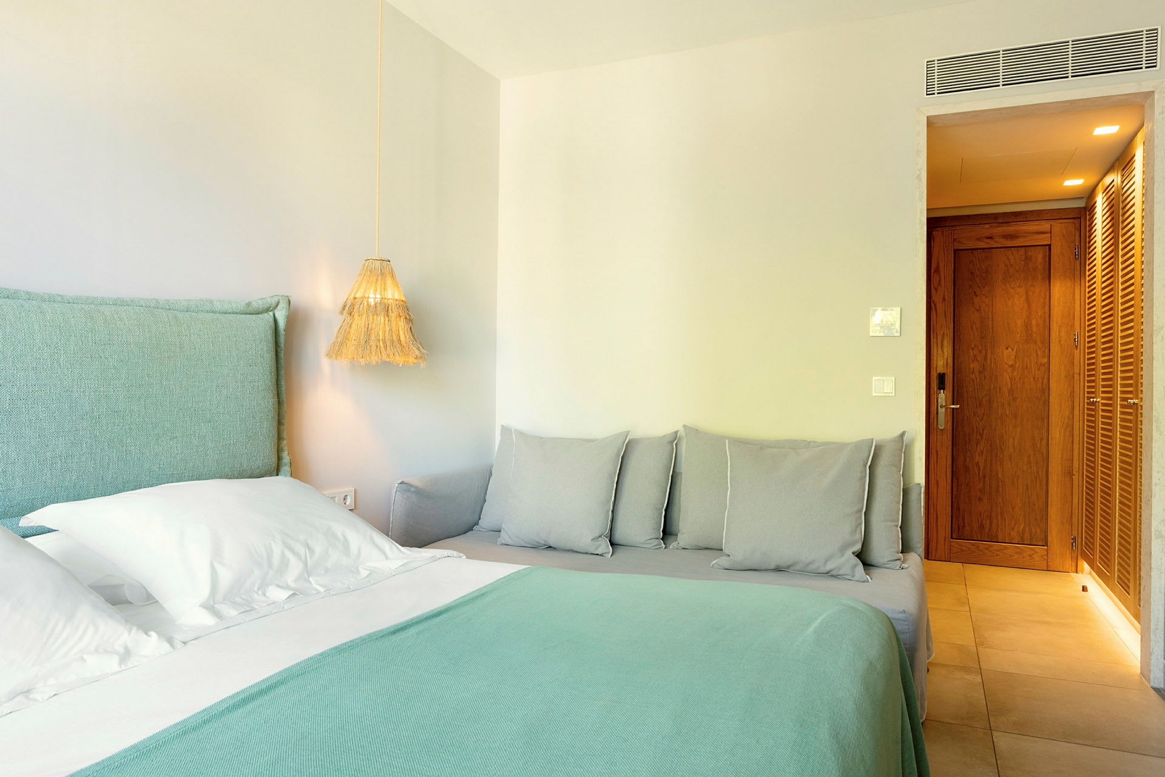 Hotellihuone, jossa sänky turkoosilla liinalla ja valkoisilla tyynyillä, sekä lähistöllä sohva harmailla tyynyillä. (Tekoälyn luoma teksti)