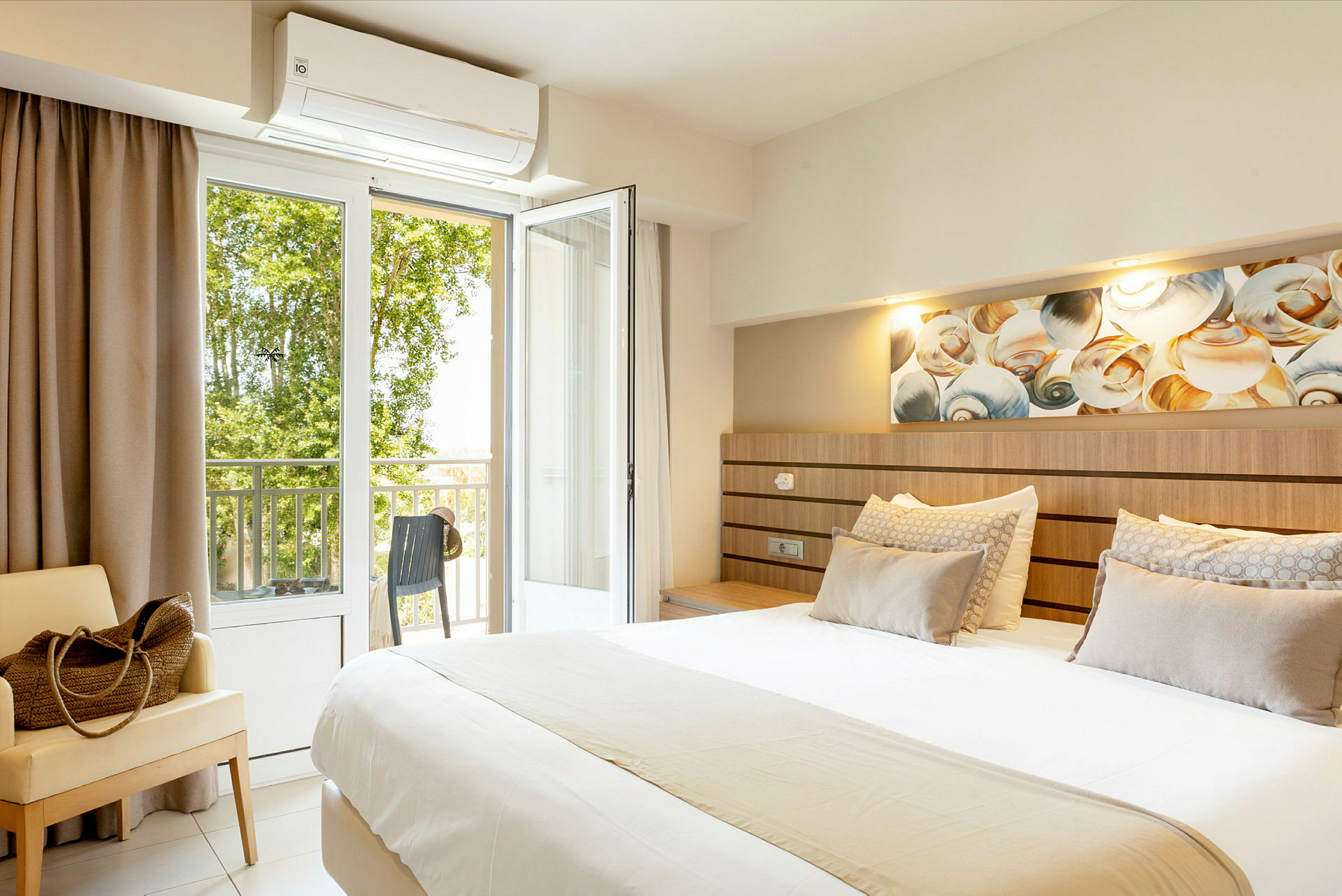 Hotellihuone, jossa suuri sänky, istuinkaluste ja avoin parvekkeen ovi näkymällä vehreyteen. (Tekoälyn luoma teksti)