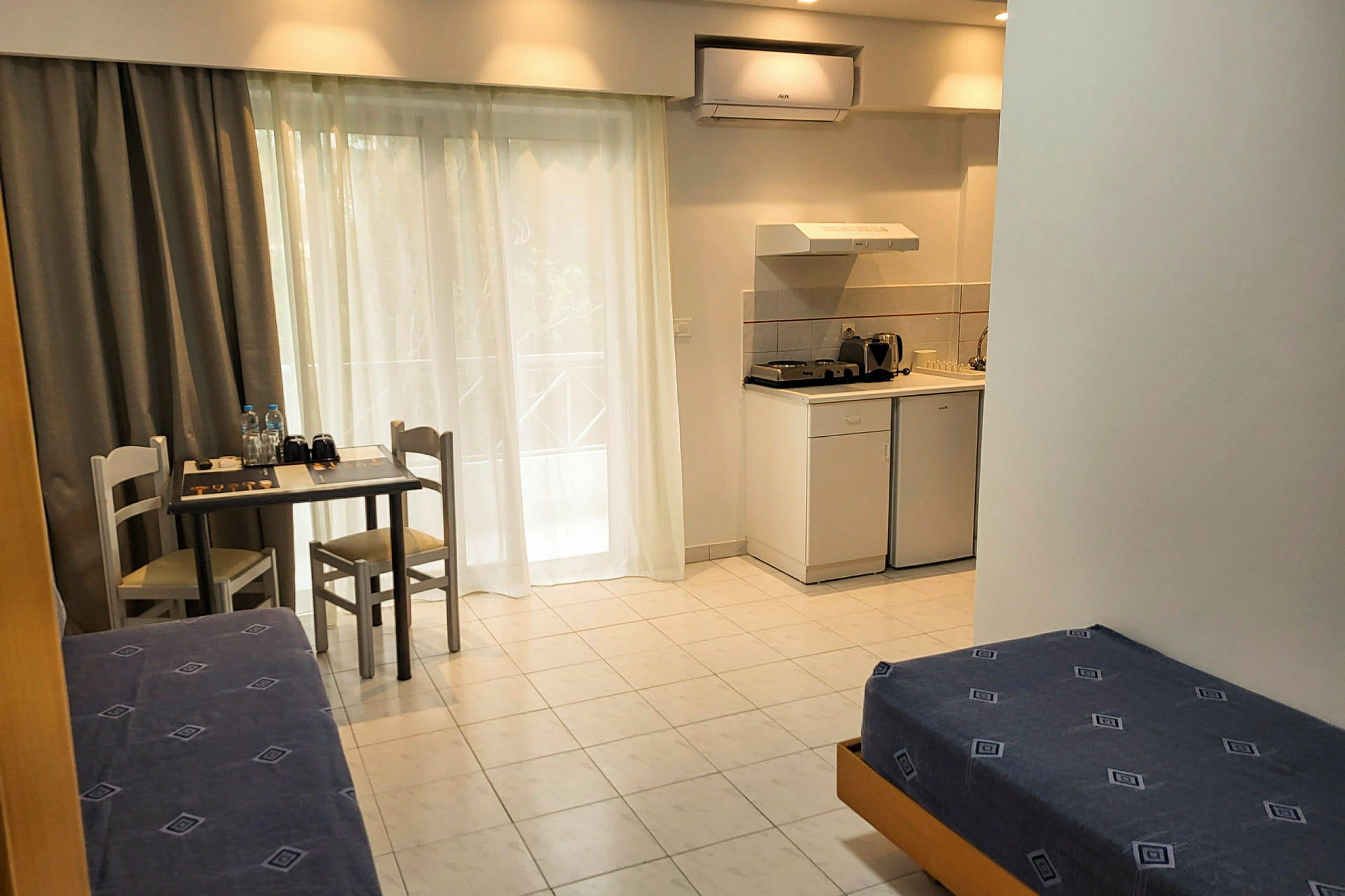 Hotellihuone, jossa on yksinkertaiset kalusteet, kuten sänkyjä, pieni pöytä ja kaksi tuolia sekä keittonurkkaus, jossa jääkaappi, liesi ja keittiövälineitä. (Tekoälyn luoma teksti)