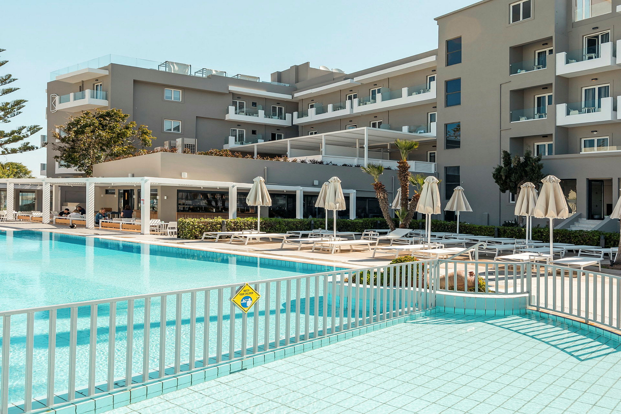 Hotellirakennus, jonka uima-allasalueella on aurinkotuoleja ja varjoja. (Tekoälyn luoma teksti)