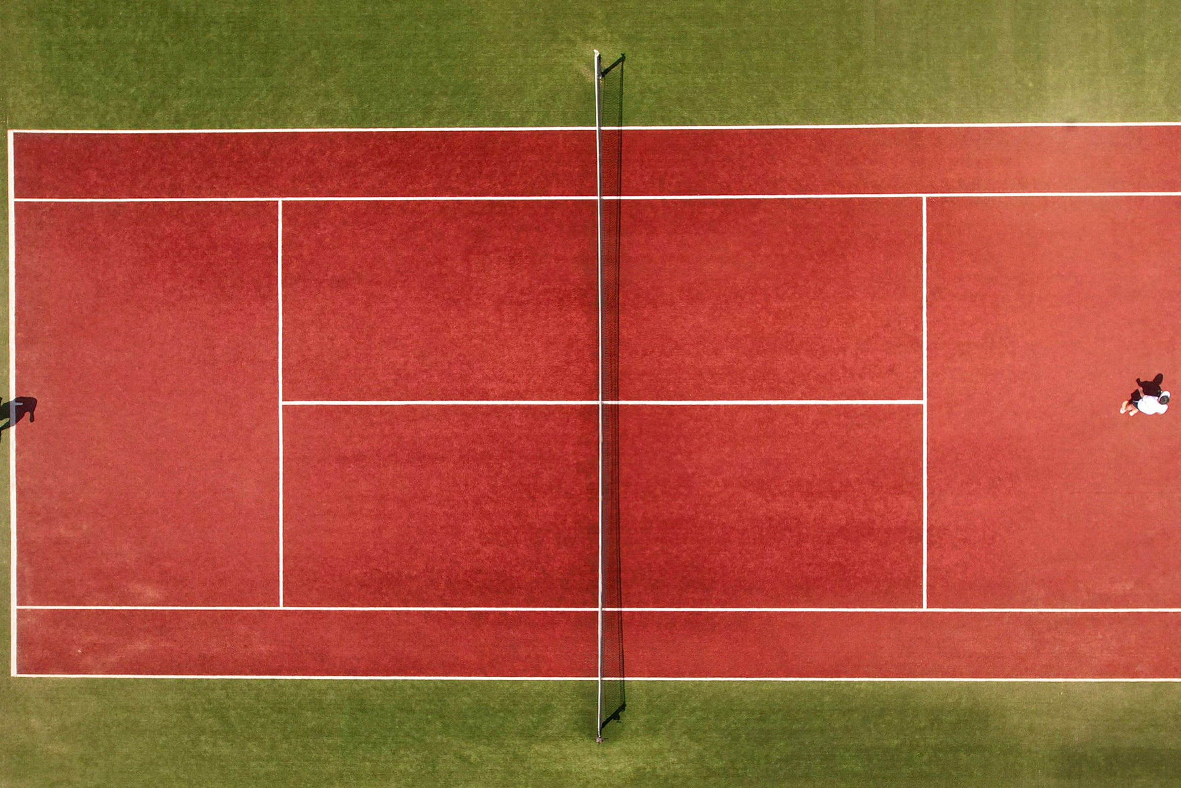 Ilmakuva punaisesta tenniskentästä verkkoineen, kaksi henkilöä pelaamassa. (Tekoälyn luoma teksti)
