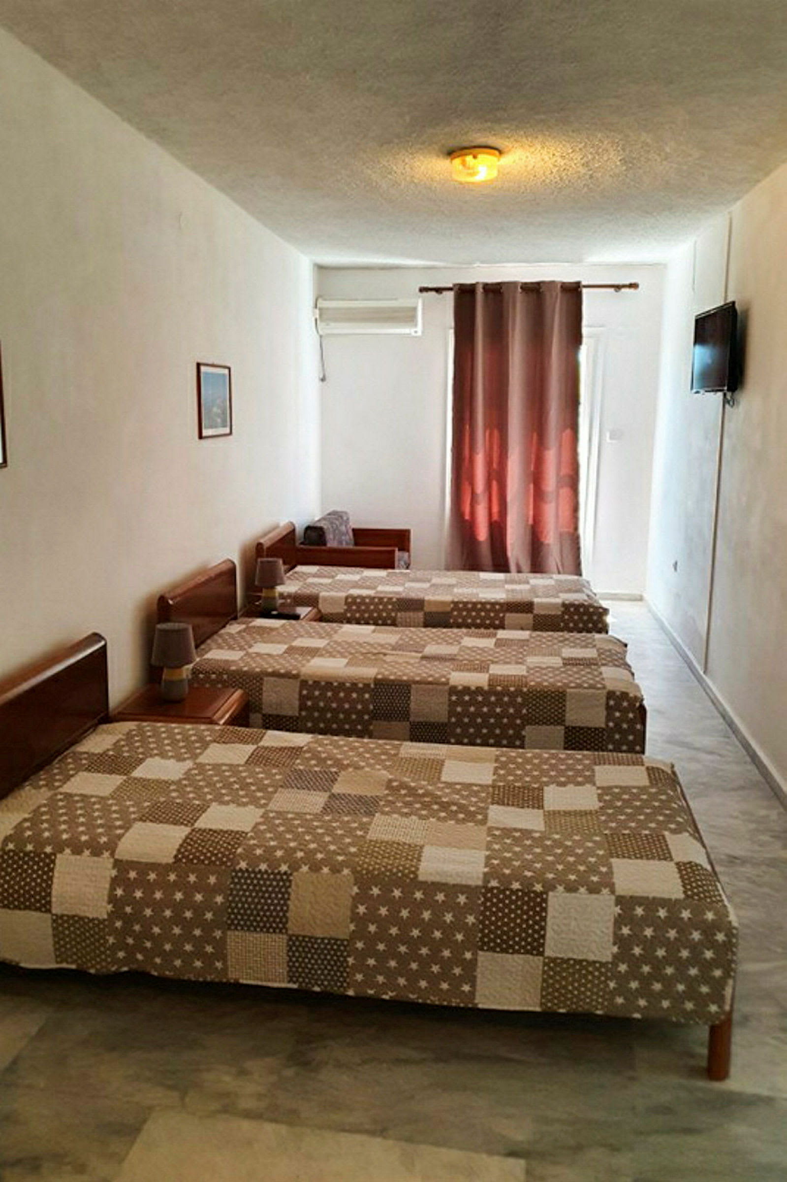 Kolme yhden hengen sänkyä hotellihuoneessa, joissa ruskeakuvioiset päiväpeitot, ilmastointi ja televisio seinällä. (Tekoälyn luoma teksti)