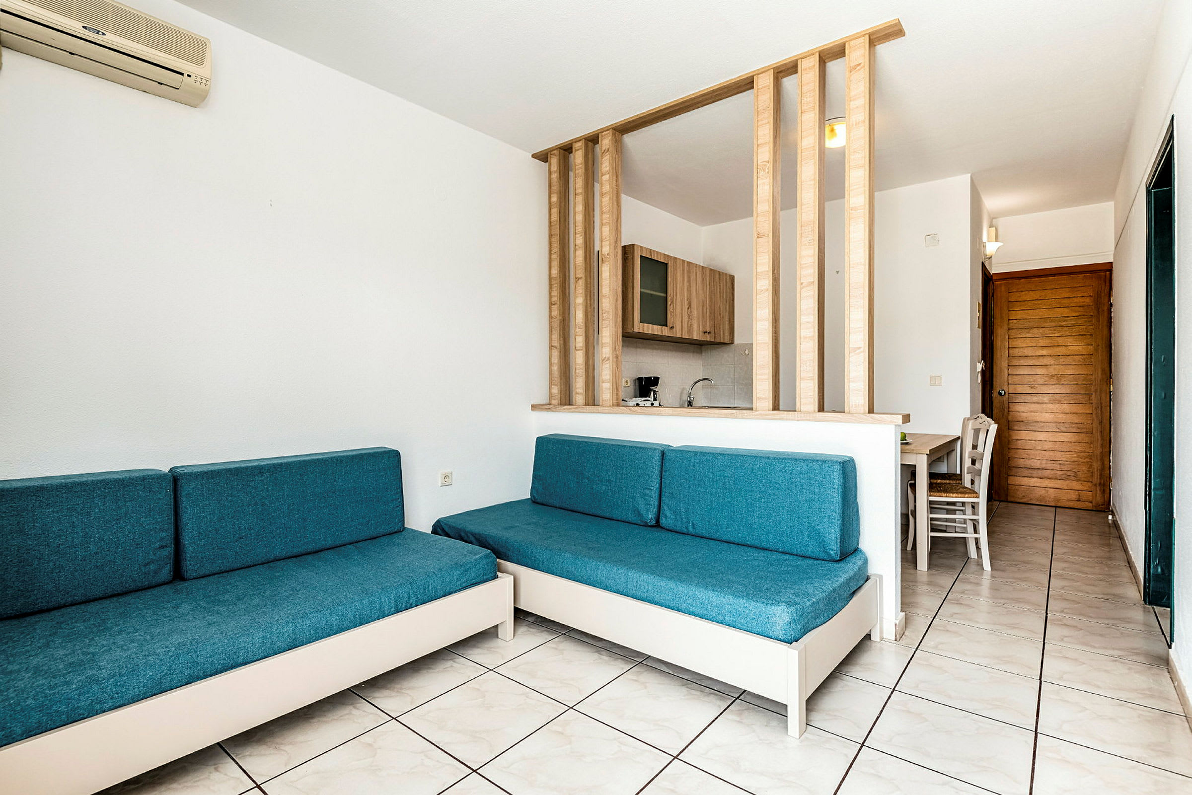 Hotellihuone, jossa on kaksi sinistä sohvaa, keittiöosio puumateriaaleilla ja ruokailutila. (Tekoälyn luoma teksti)