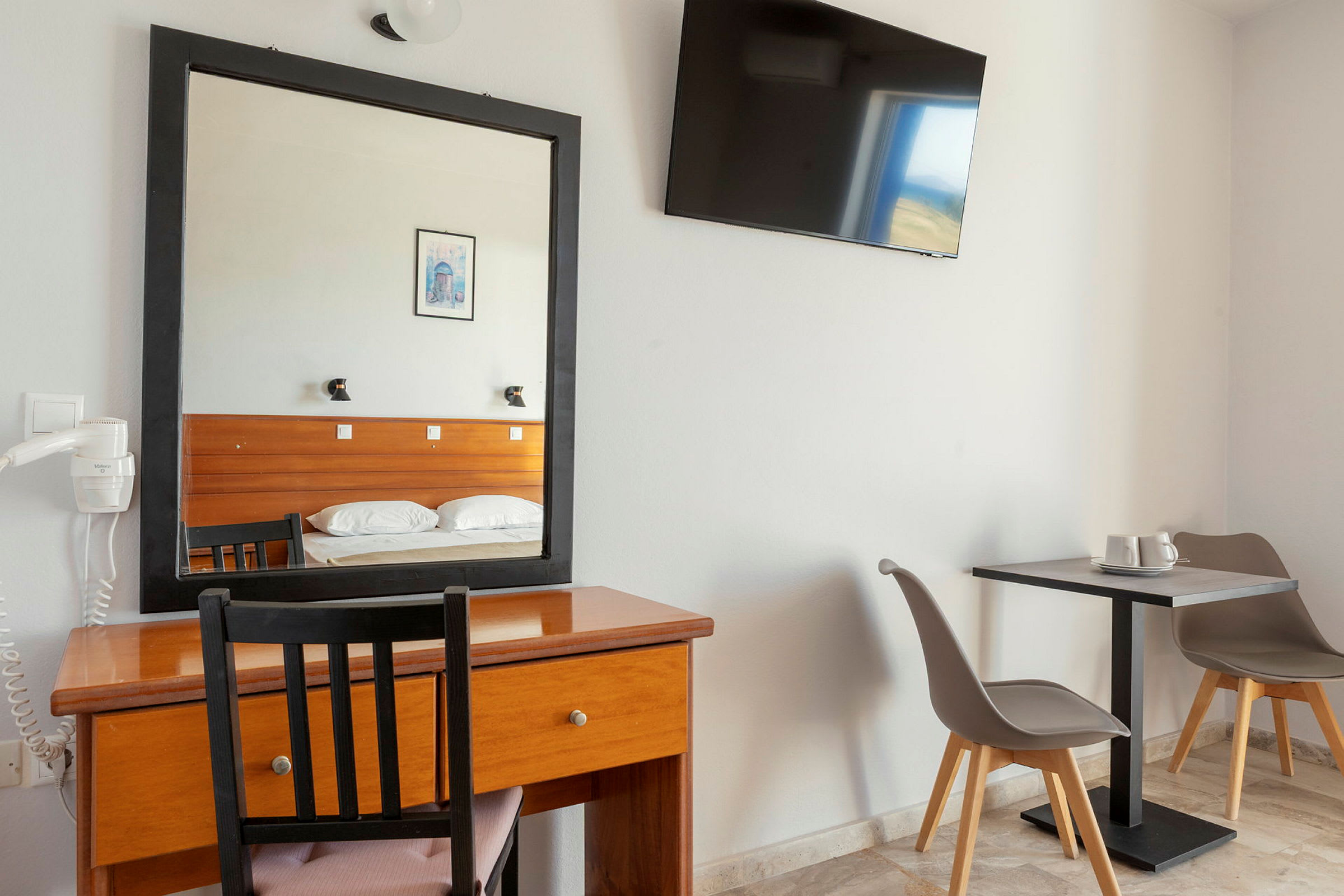 Hotellihuone, jossa kirjoituspöytä, peili ja televisio seinällä. Oikealla kaksi tuolia pöydän ääressä. (Tekoälyn luoma teksti)