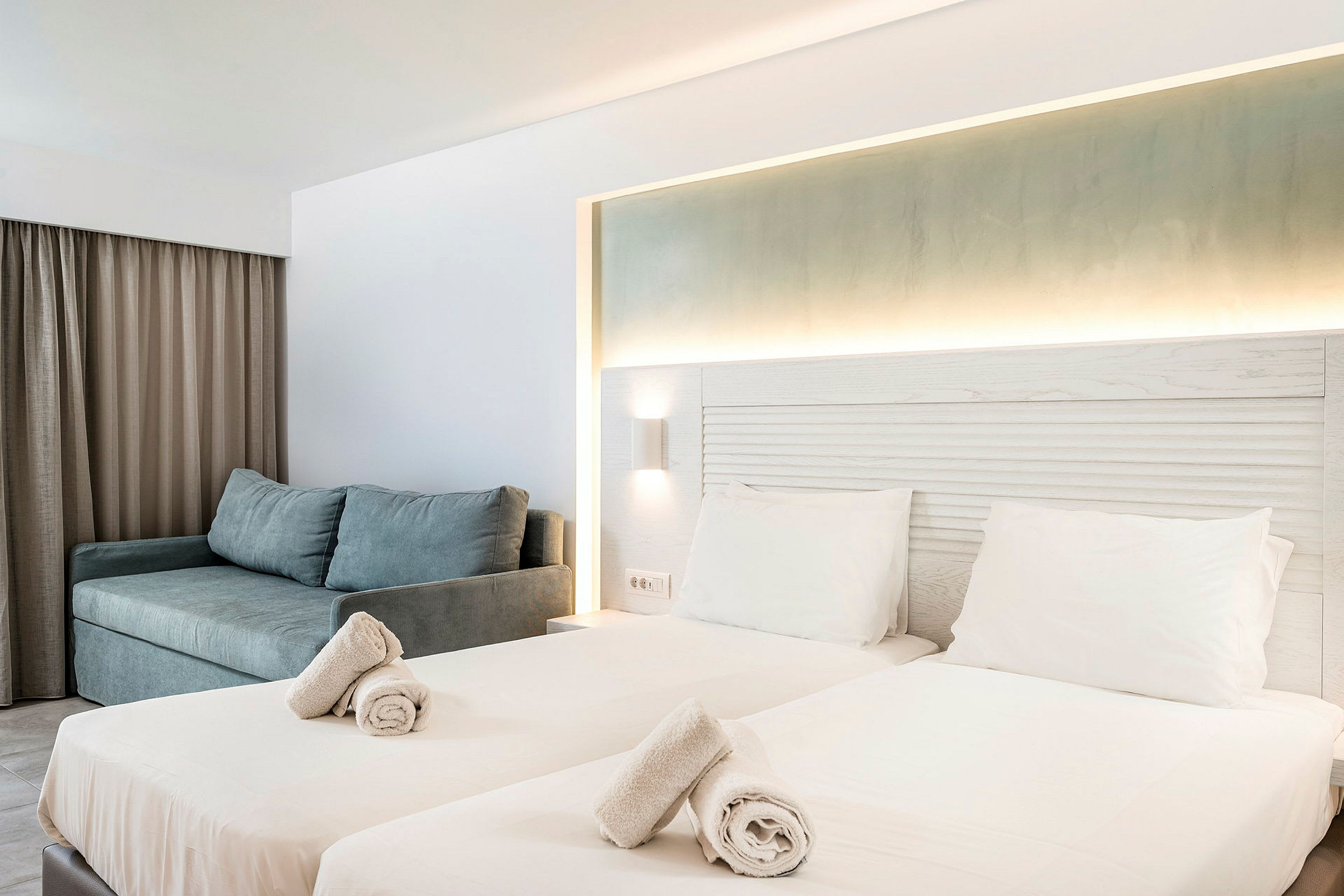 Hotellihuone, jossa kaksi erillistä vuodetta, valaistu sängynpääty ja sohva. (Tekoälyn luoma teksti)