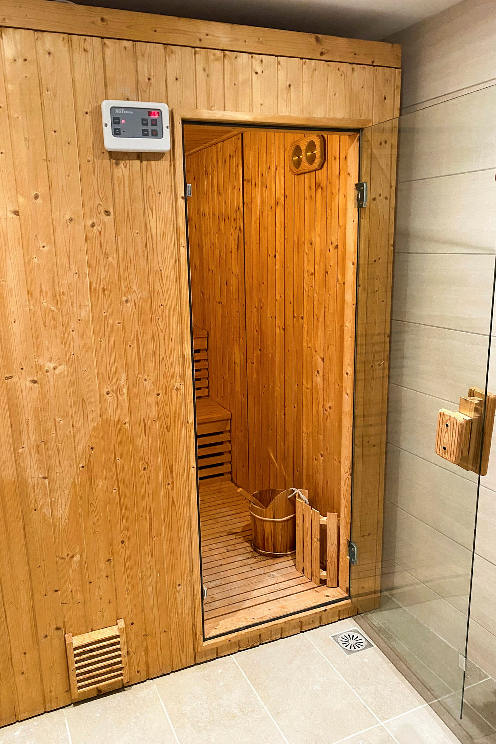 Puutuinen sisäsauna, jossa avoin ovi, puuämpäri ja lattiakaivo näkyvissä. (Tekoälyn luoma teksti)