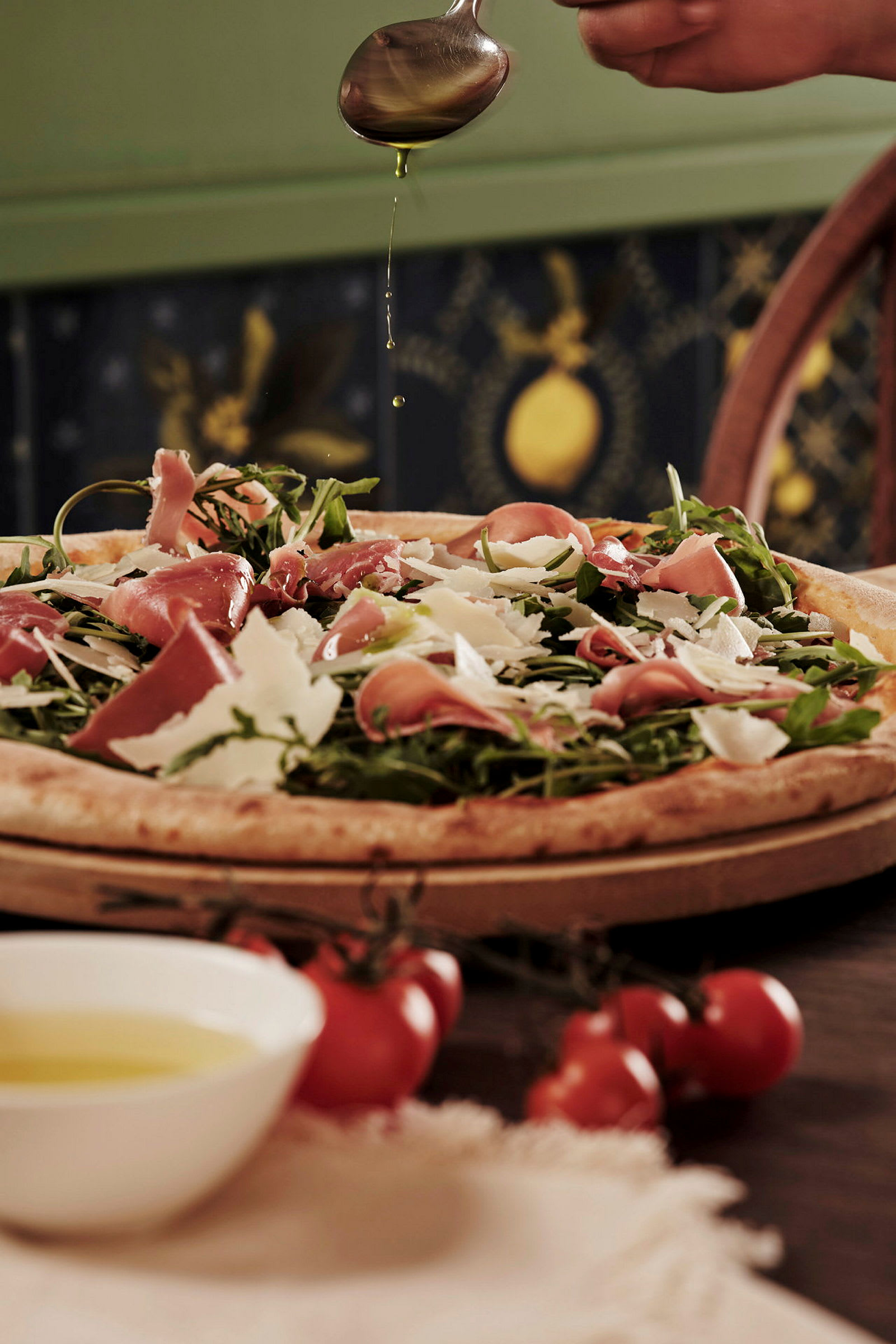 Pizza, jonka päällä on kinkkua, rucolaa ja parmesania, käsi tiputtaa päälle oliiviöljyä. (Tekoälyn luoma teksti)