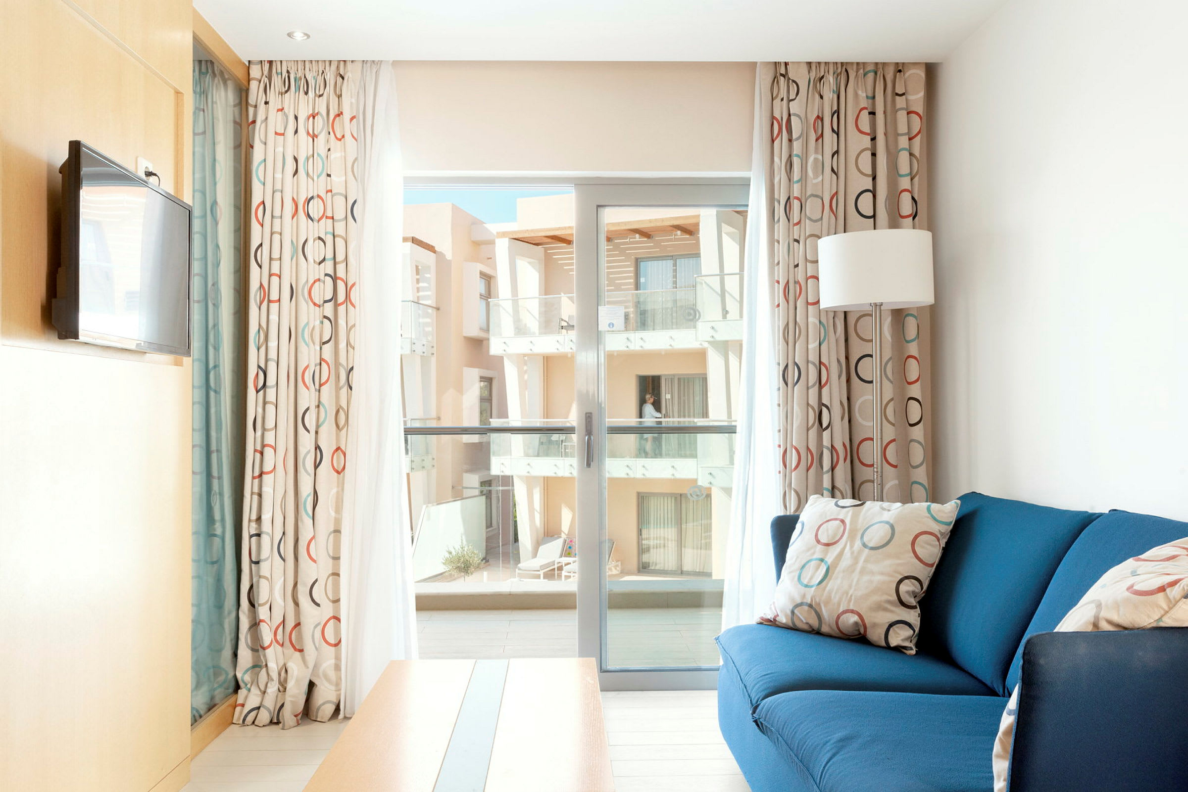 Valoisa hotellihuone, jossa on sininen sohva, ympyräkuvioiset verhot ja parveke käynti. (Tekoälyn luoma teksti)