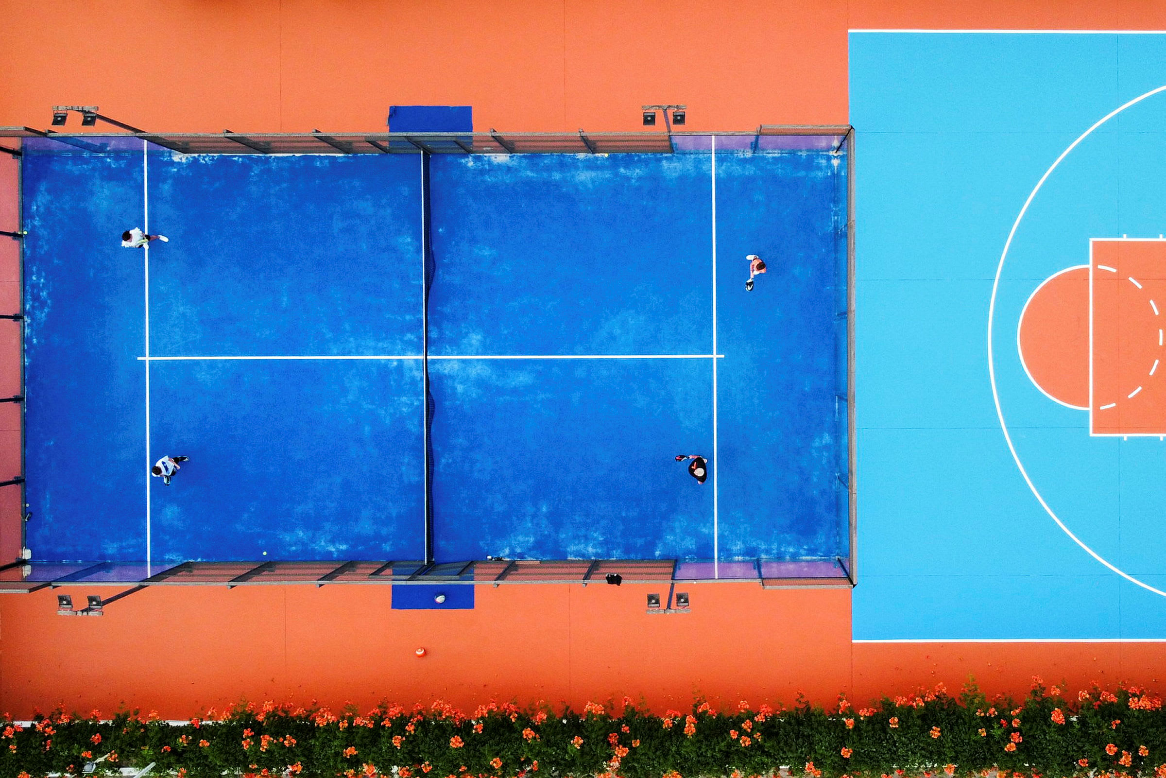 Ilmakuva sinisestä padelkentästä, jossa ihmiset pelaavat, viereisen oranssi-sinisen koripallokentän ohella. (Tekoälyn luoma teksti)