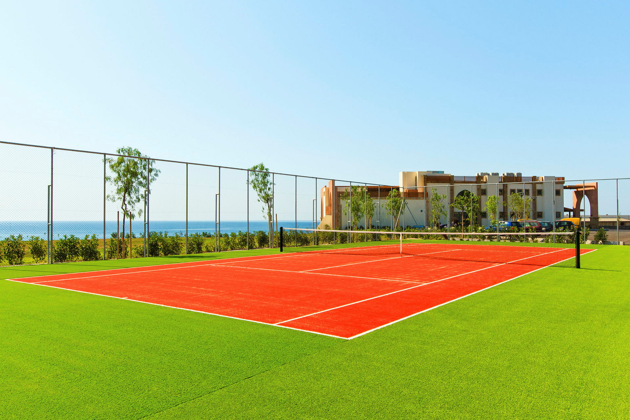 Punaisella pinnalla varustettu tenniskenttä, jota ympäröi vihreä nurmikko, taustalla meri ja rakennus. (Tekoälyn luoma teksti)