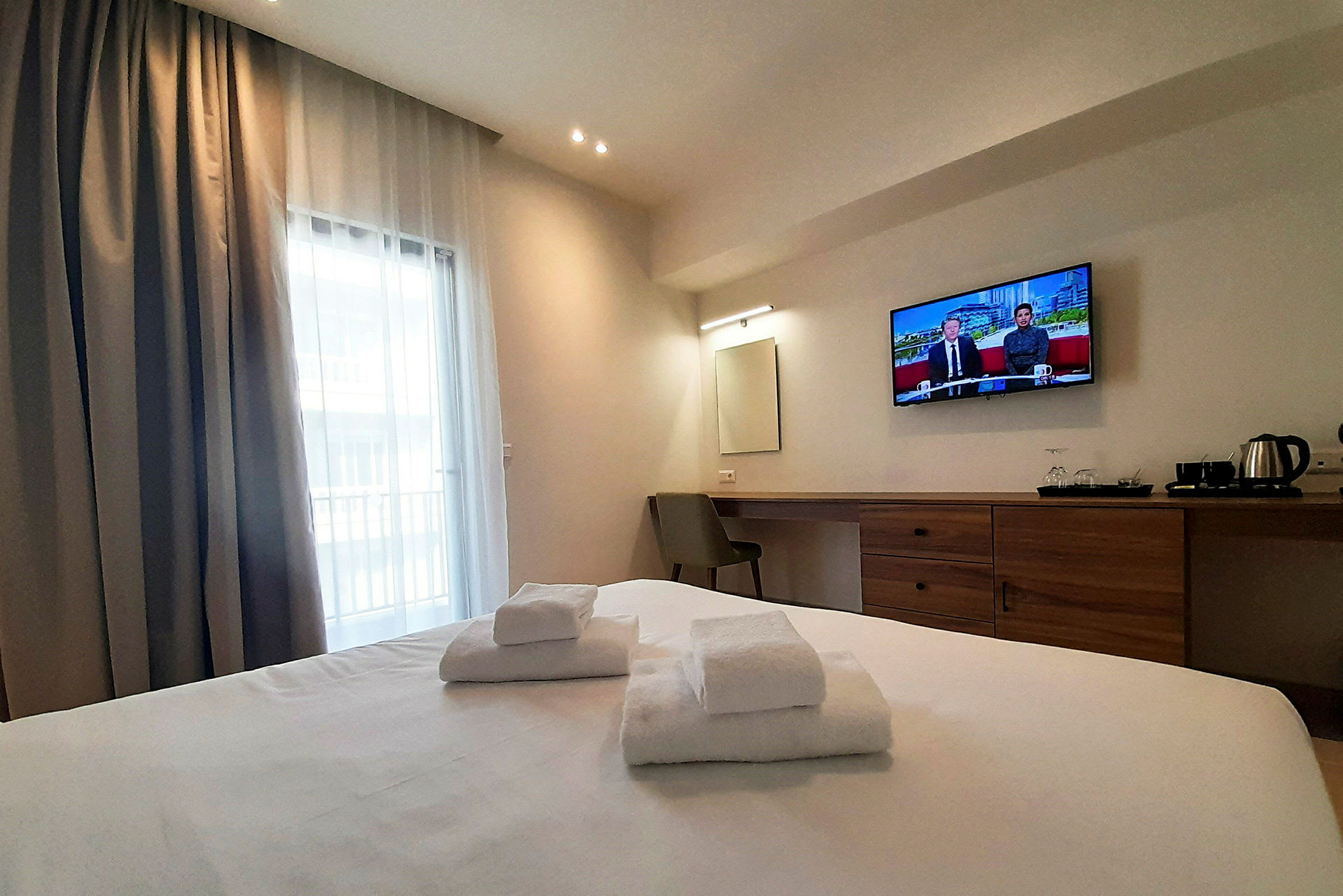 Hotellihuone, jossa on parivuode, valkoiset pyyhkeet, seinään kiinnitetty televisio ja työpöytä tuolilla. Ikkuna, jossa on verhot, näkyy. (Tekoälyn luoma teksti)