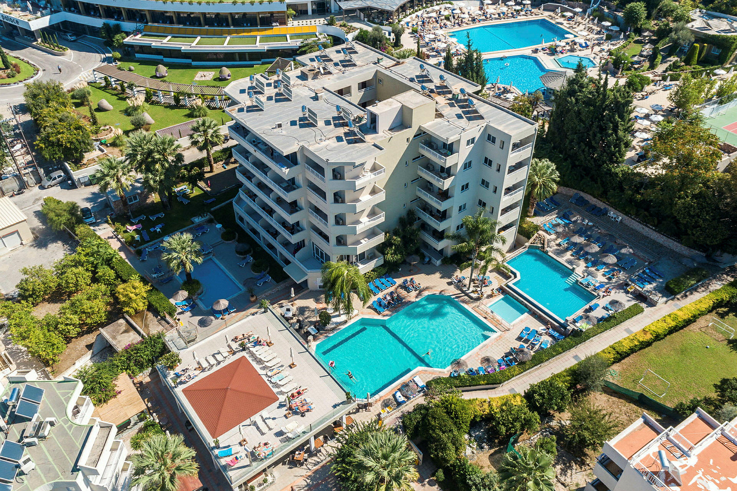 Ilmakuva hotellista uima-allasalueiden, aurinkotuolien ja vehreyden kera. (Tekoälyn luoma teksti)