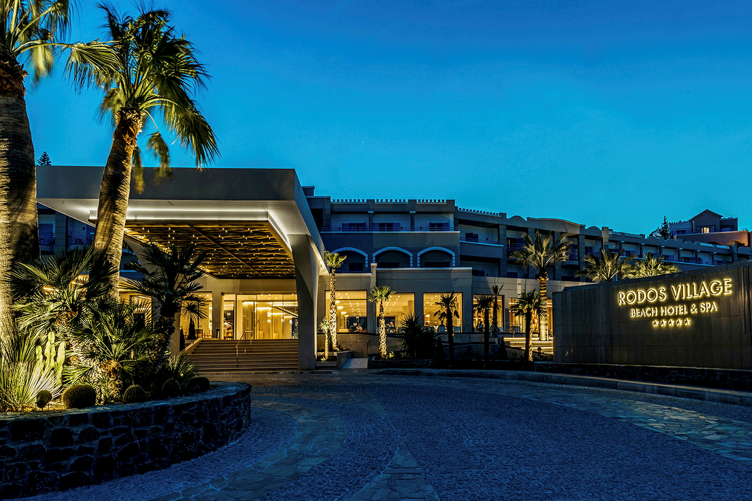 Hotelli illalla, jossa valaistu sisäänkäynti ja kyltti, jossa lukee 'Rodos Village Beach Hotel & Spa'. (Tekoälyn luoma teksti)