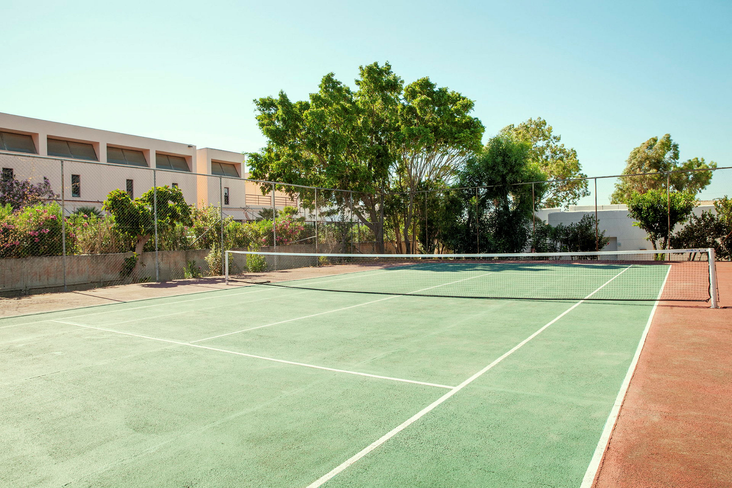 Tenniskenttä, jossa on vihreä pinta ja verkko, ympäröitynä puilla ja rakennuksilla. (Tekoälyn luoma teksti)