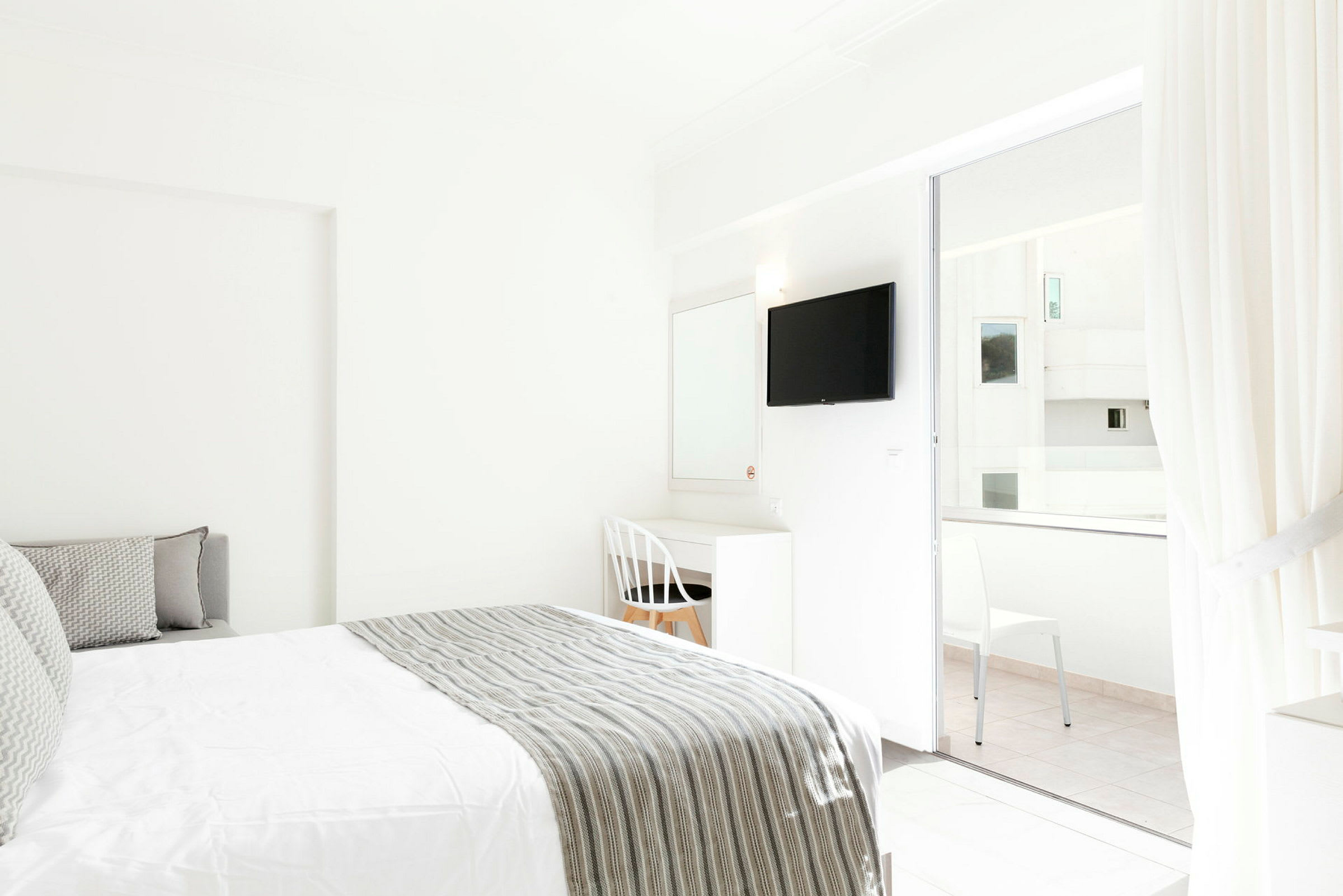 Valoisan hotellihuoneen sisustus, jossa on sänky ja kirjoituspöytä valkoisen tuolin vieressä. TV on kiinnitetty seinään, ja lasiovet vievät parvekkeelle. (Tekoälyn luoma teksti)