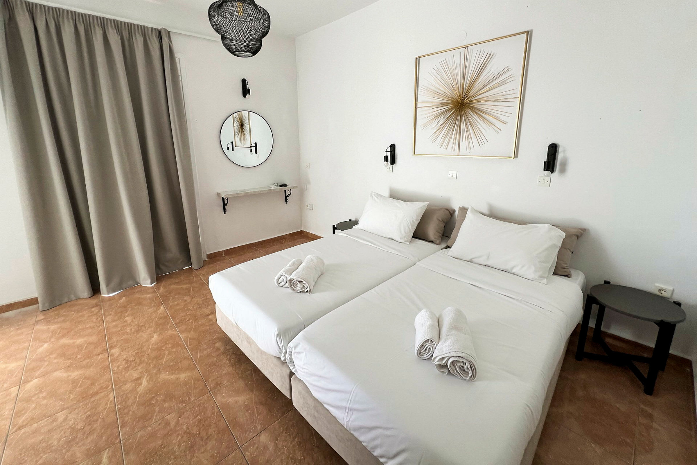 Kaksi yhden hengen sänkyä, joissa on valkoiset vuodevaatteet ja taitetut pyyhkeet päällä, ovat hotellihuoneessa. Pyöreä peili ja seinäkoriste näkyvät seinällä. (Tekoälyn luoma teksti)