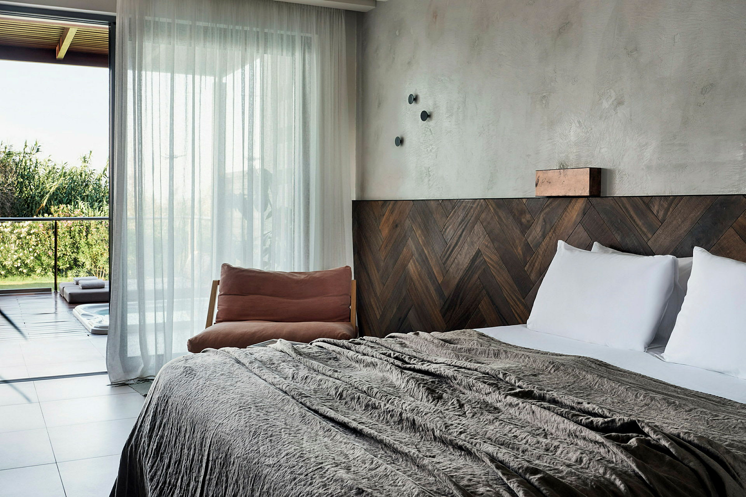 Hotellihuone, jossa on iso sänky, puukoristeita ja tuoli vieressä, sekä lasiovet, jotka johtavat terassille, josta on näkymä vihreään ympäristöön. (Tekoälyn luoma teksti)