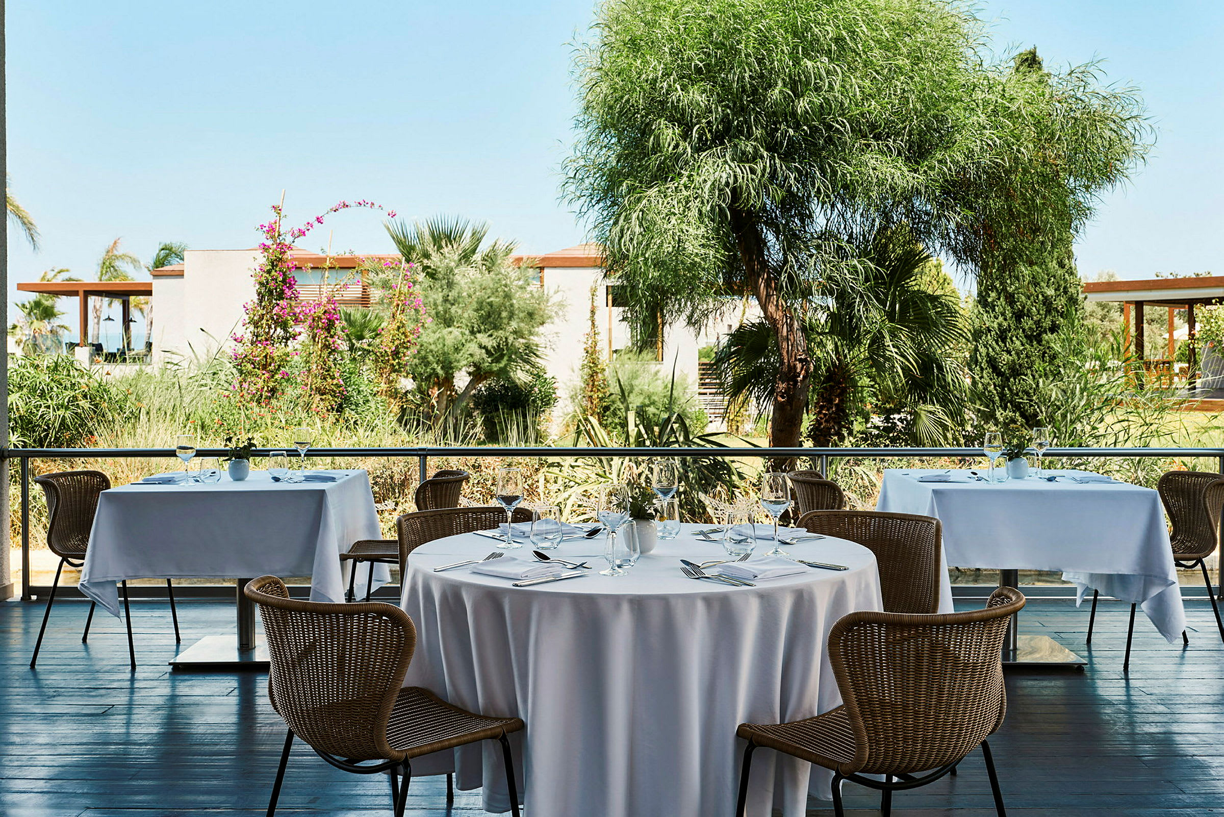 Pöydät, joissa on valkoiset liinat ja tuolit, terassilla näkymä vehreään puutarhaan. (Tekoälyn luoma teksti)
