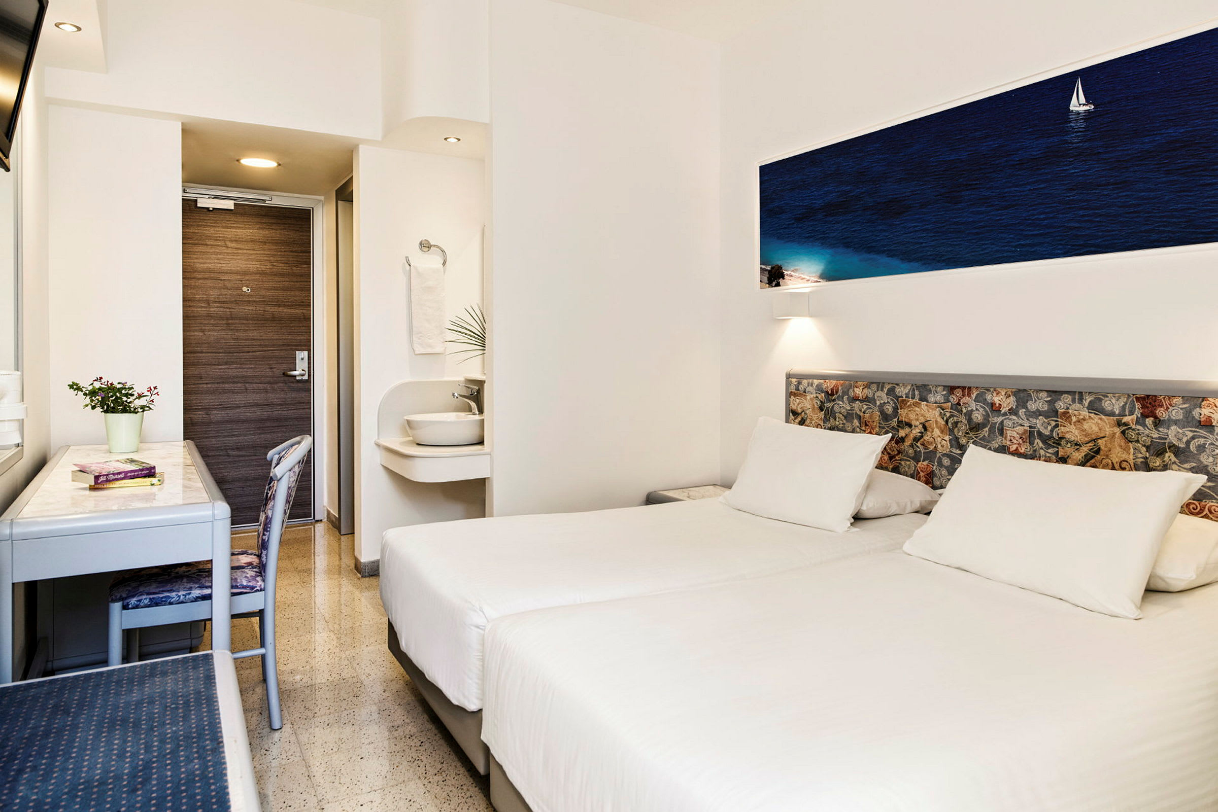 Hotellihuone, jossa on kaksi valkoista yhden hengen vuodetta, kirjoituspöytä, tuoli ja pieni pesuallas kylpyhuoneen ulkopuolella. (Tekoälyn luoma teksti)