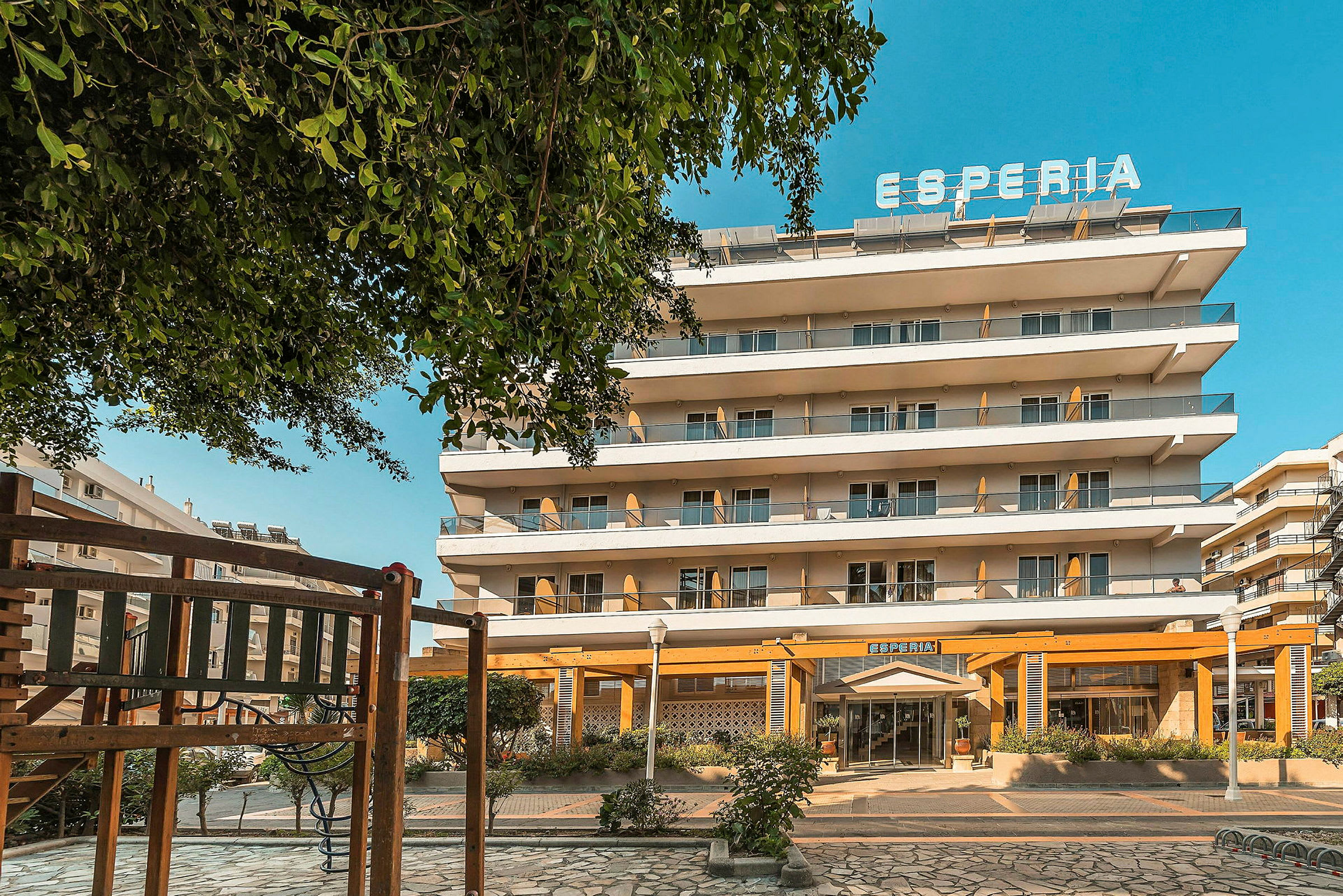 Hotellirakennus, jossa on useita kerroksia ja ylhäällä kyltti, jossa lukee 'Esperia'. Etualalla on leikkivälineitä. (Tekoälyn luoma teksti)