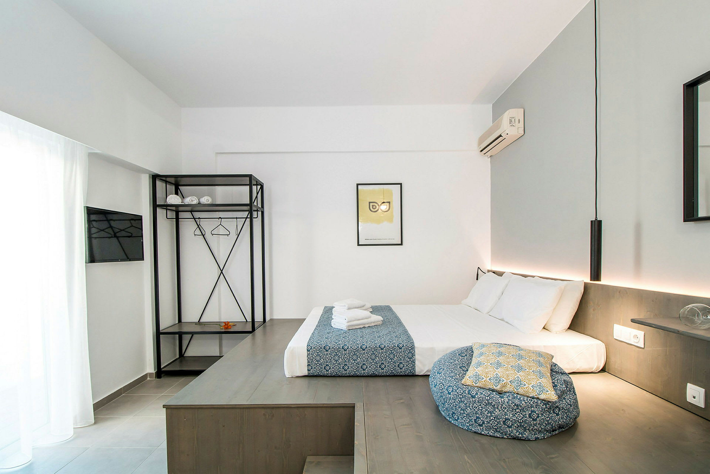 Moderni hotellihuone, jossa sänky, valkoiset ja siniset tekstiilit, avoin vaatekaappi, hyllyt ja vaateripustimet sekä seinään kiinnitetty ilmastointilaite. (Tekoälyn luoma teksti)