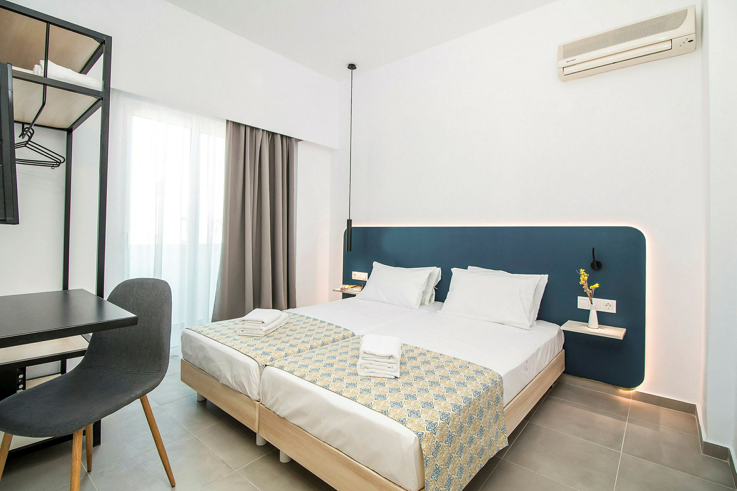 Moderni hotellihuone, jossa kaksi pedattua yhden hengen vuodetta, kirjoituspöytä ja tuoli sekä seinään kiinnitetty ilmastointilaite. (Tekoälyn luoma teksti)
