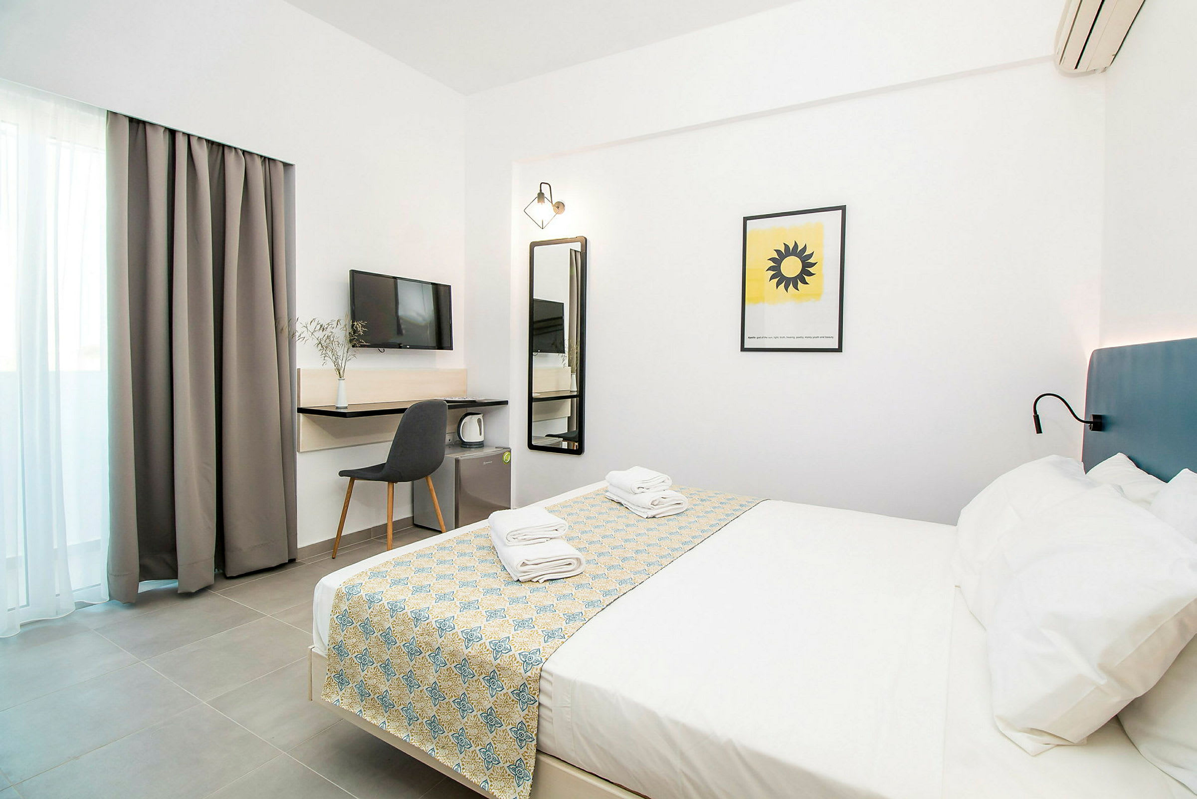 Hotellihuone, jossa on valkoiset sisätilat, parisänky, johon on pedattu kuviollinen päiväpeite ja pyyhkeet sekä huonekalut ja televisio. (Tekoälyn luoma teksti)