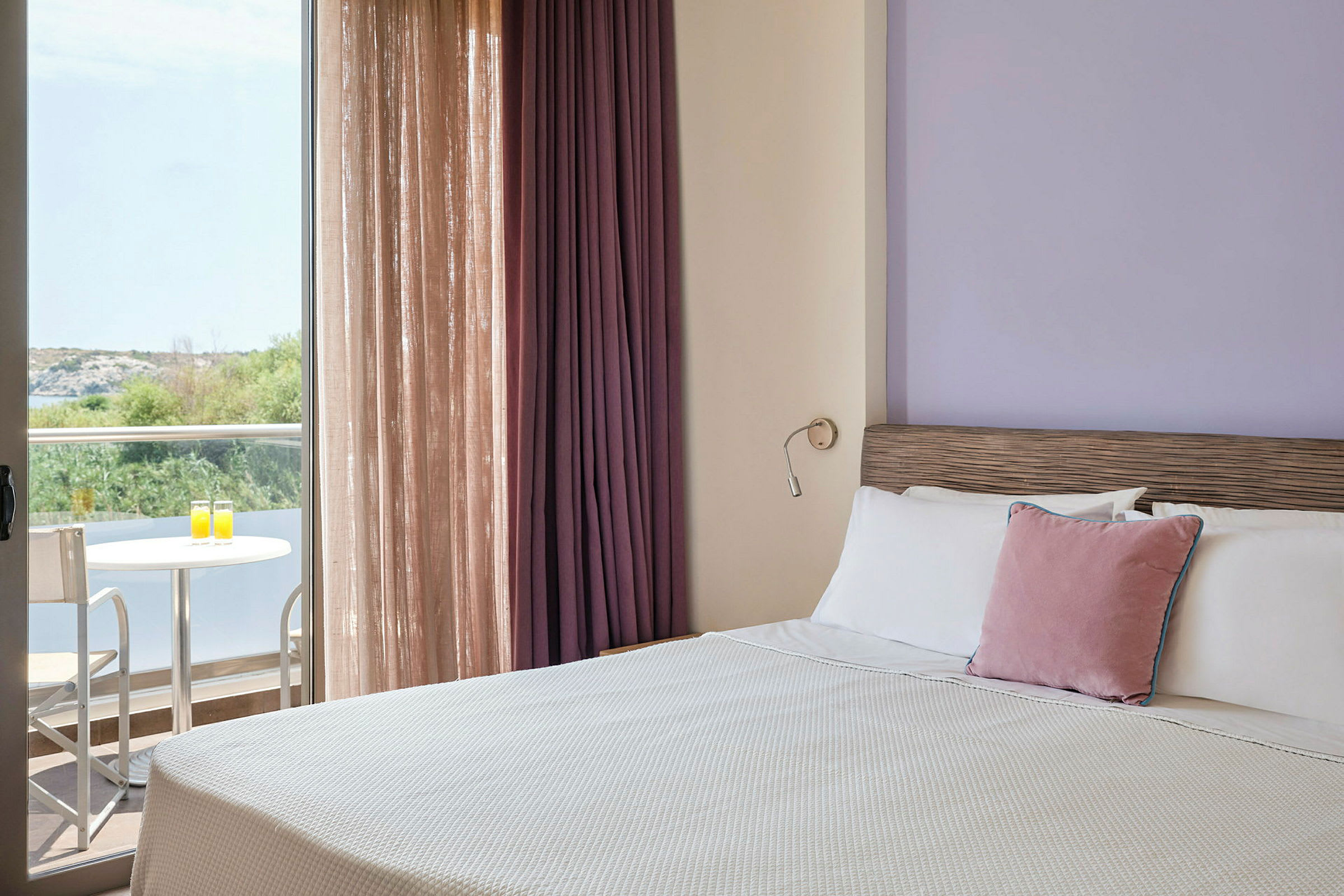 Hotellihuone, jossa on pedattu sänky, violetti seinä ja verhot sekä parveke, jossa pöytä ja kaksi tuolia, näkymä vehreyteen. (Tekoälyn luoma teksti)