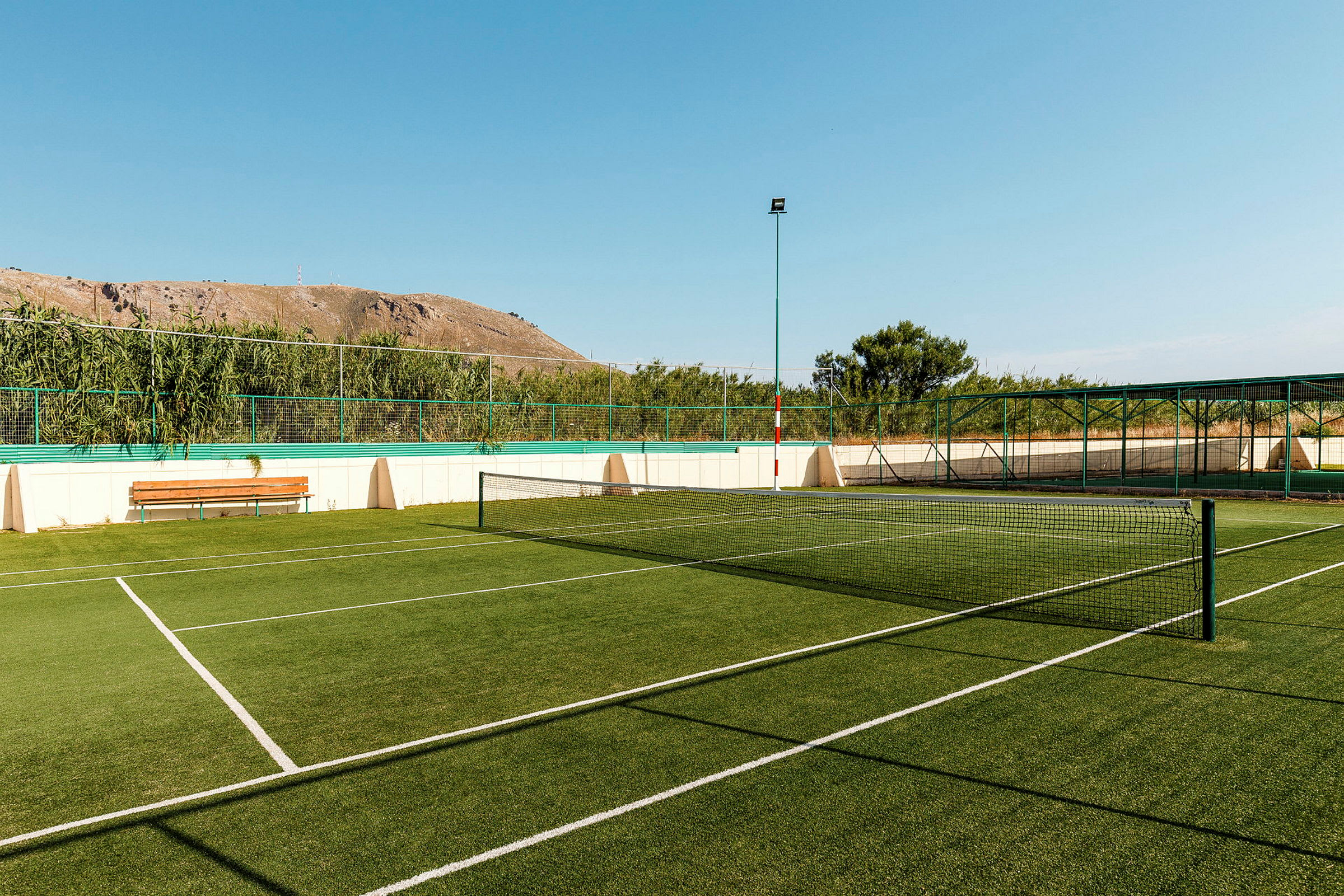 Tennis-kenttä vihreällä tekonurmella, jota ympäröivät aidat ja kasvillisuus. (Tekoälyn luoma teksti)