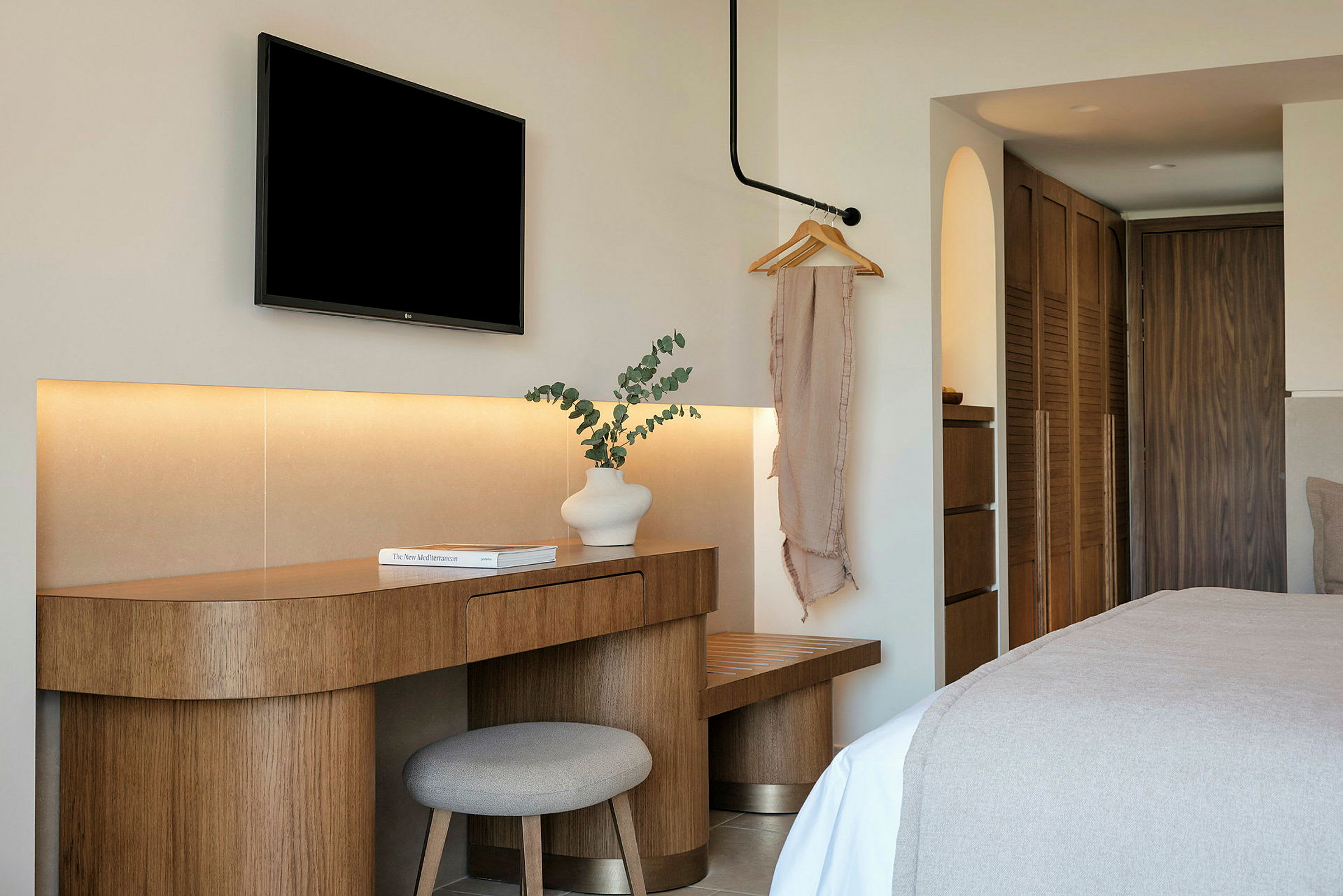 Hotellihuone, jossa on moderni sisustus. Puupöytä, valkoinen maljakko ja kirja sijaitsevat tuolin vieressä. Musta televisio on kiinnitetty seinälle yläpuolella. (Tekoälyn luoma teksti)
