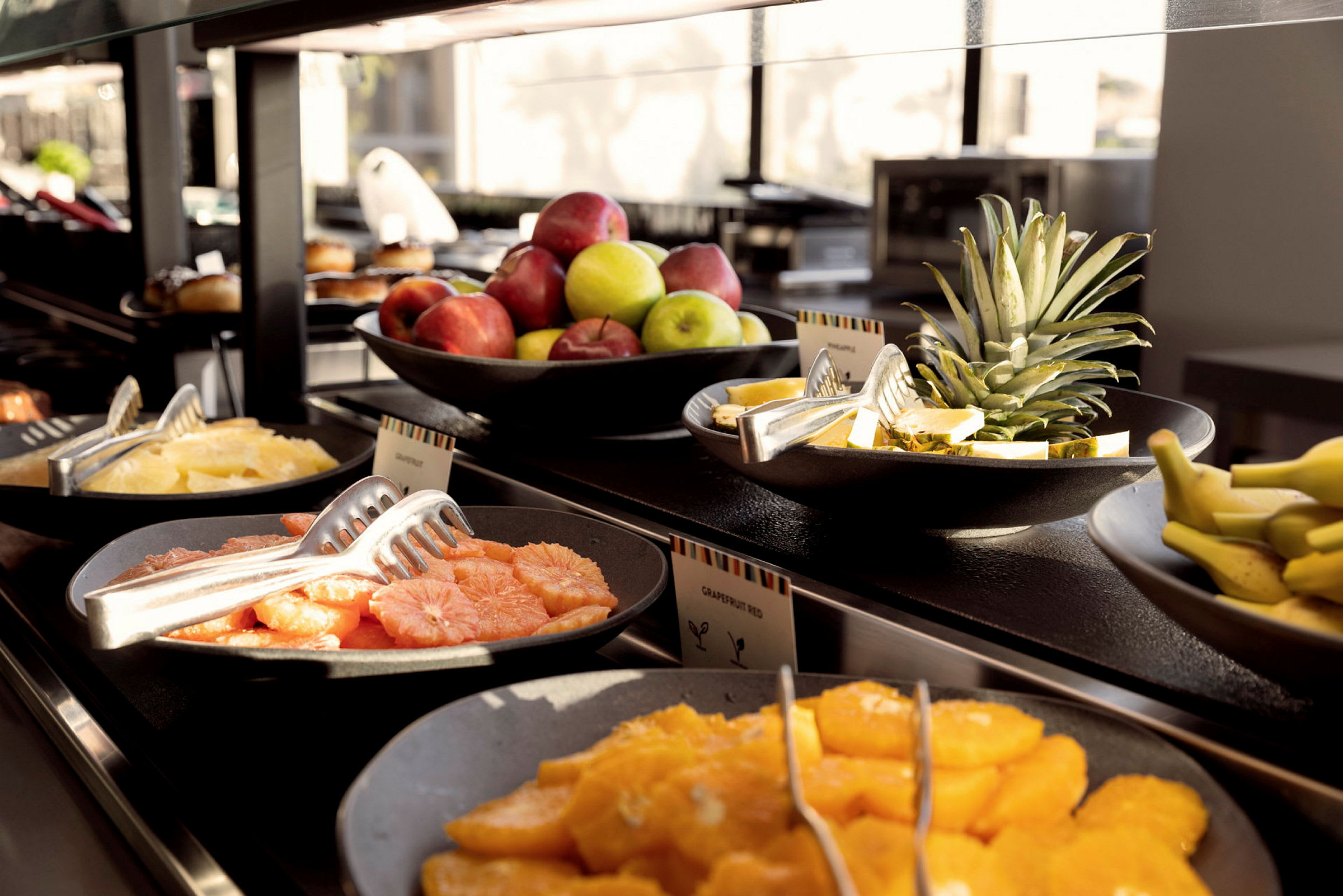 Buffet, jossa on kulhoja, jotka sisältävät tuoreita hedelmiä, kuten appelsiineja, greippejä, omenoita, banaaneja ja ananasta. (Tekoälyn luoma teksti)