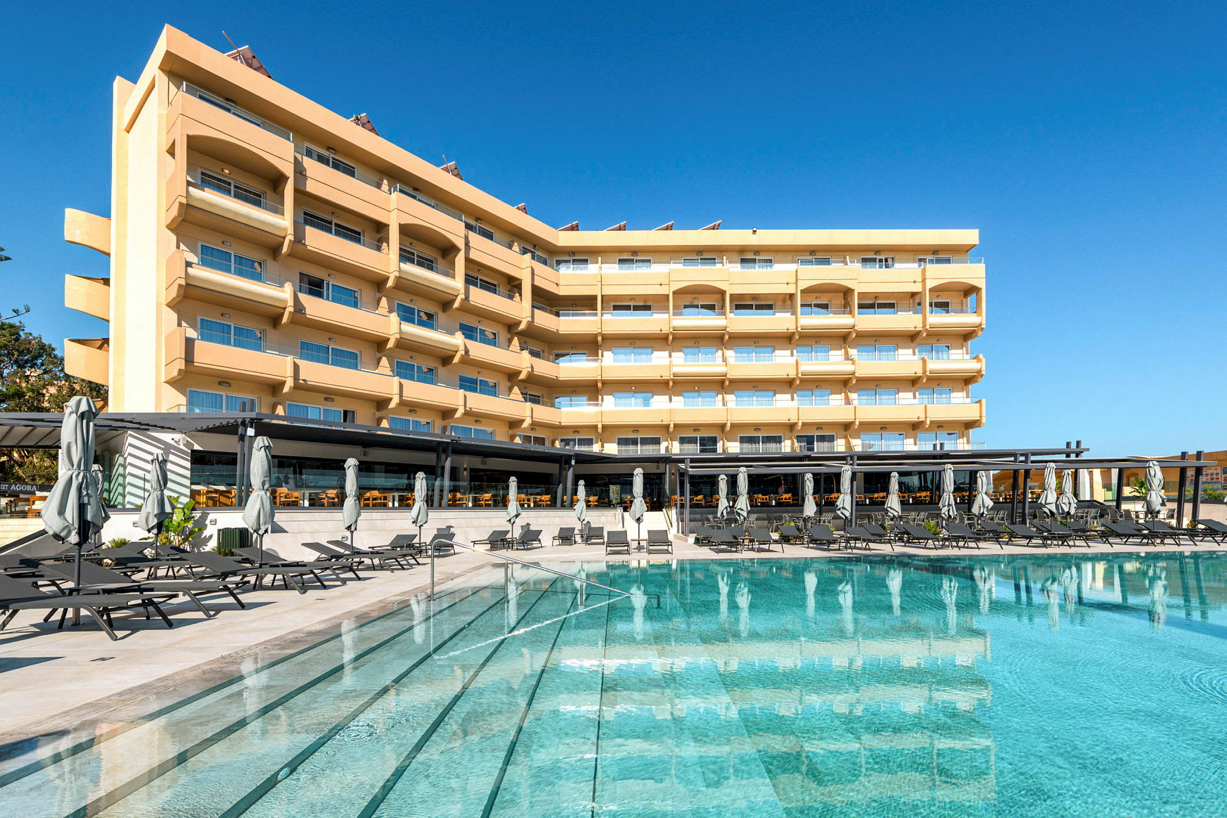 Hotellirakennus, jossa useita kerroksia, parvekkeita ja suuria ikkunoita, uima-altaan edessä aurinkotuoleineen ja aurinkovarjoineen. (Tekoälyn luoma teksti)