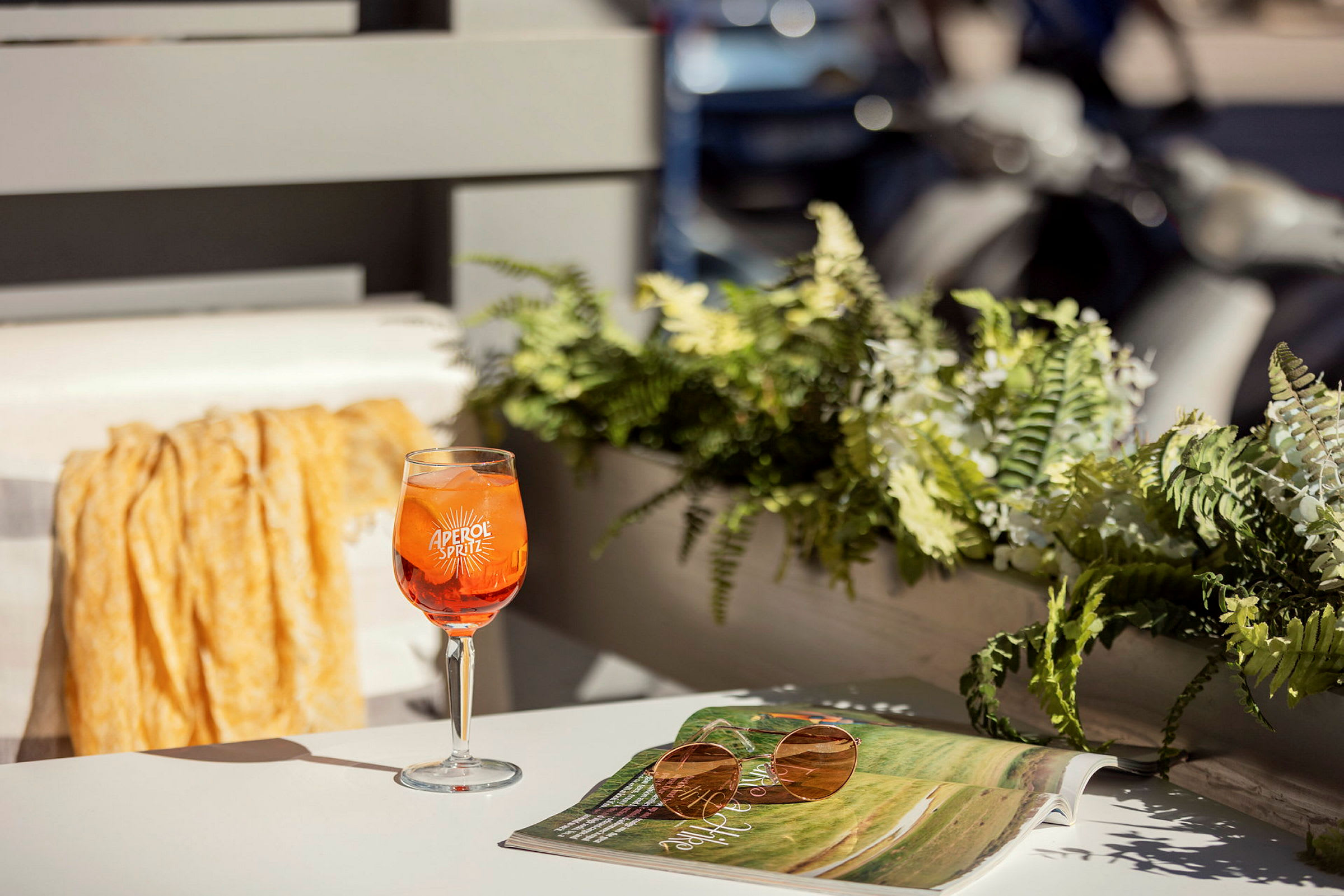 Aperol Spritz -lasillinen seisoo pöydällä aurinkolasien ja avoimen lehden vieressä. Taustalla näkyy kasveja ja parvekkeen osia. (Tekoälyn luoma teksti)