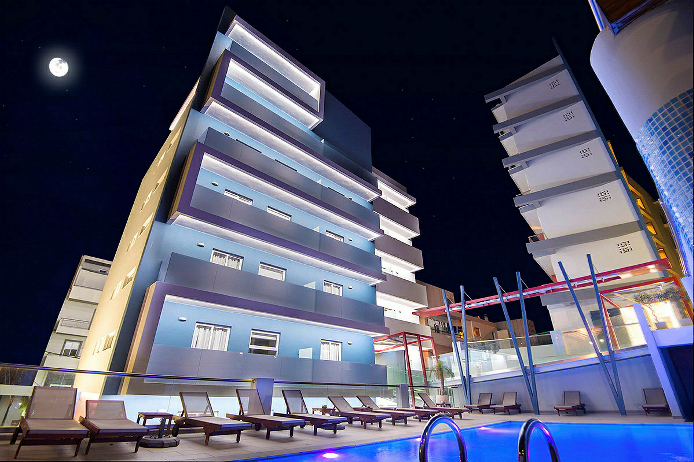Yökuva nykyaikaisesta hotellista, jossa valaistut julkisivut, uima-allas etualalla ja ympärillä aurinkotuoleja. (Tekoälyn luoma teksti)