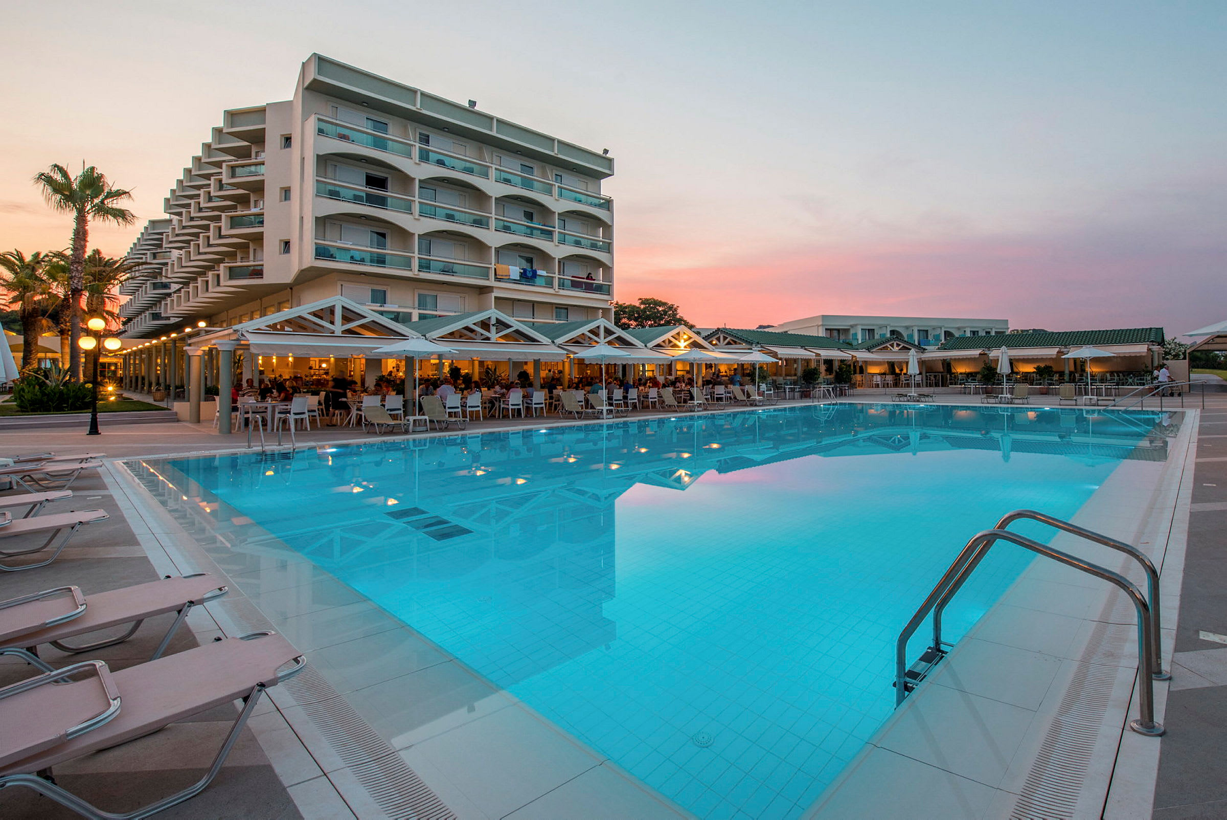 Hotellirakennus parvekkeineen suuren uima-altaan äärellä, ympärillä aurinkotuoleja ja terassi, jossa istumapaikkoja. (Tekoälyn luoma teksti)