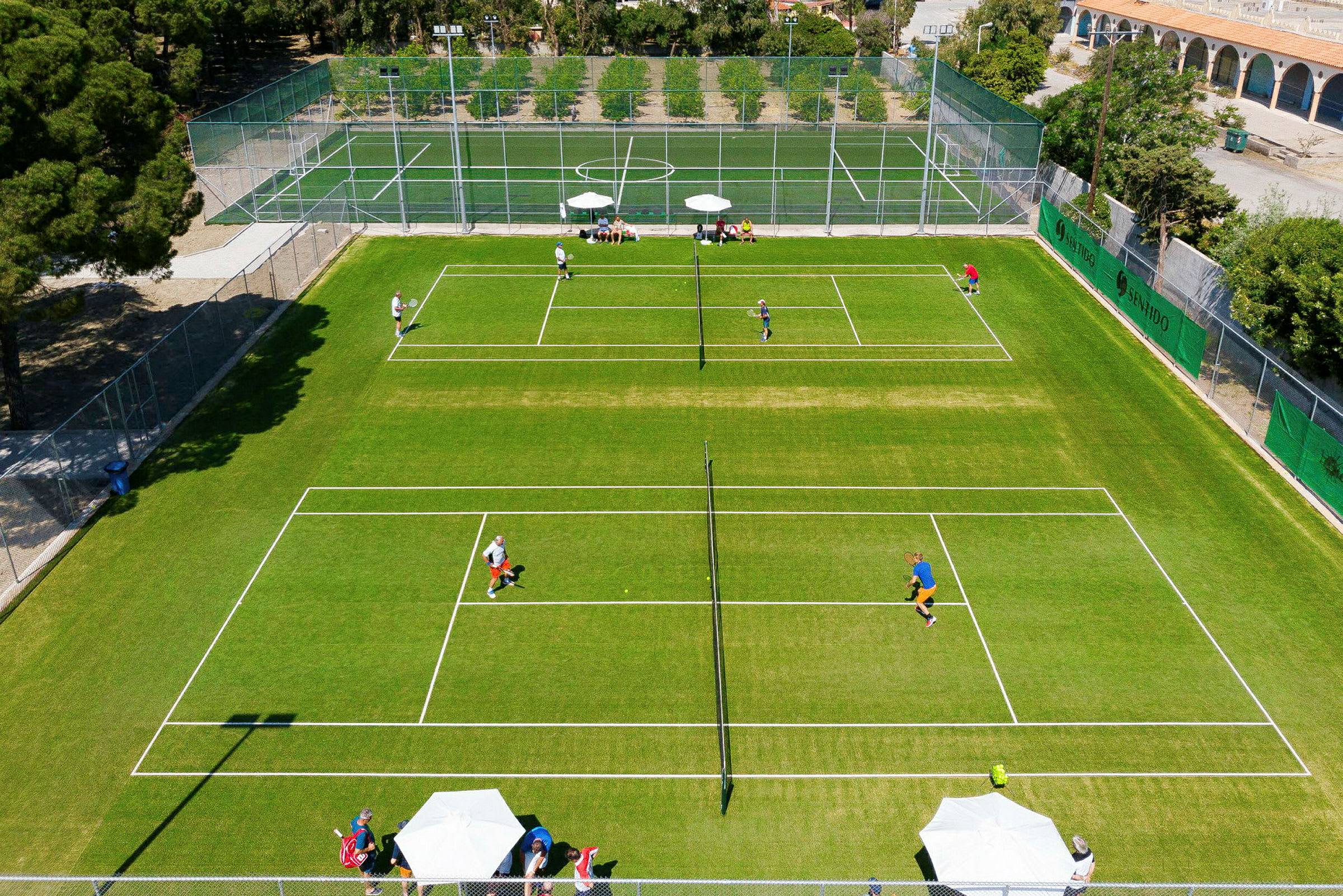 Ilmakuva tenniskentästä pelaajineen ja ympäröivistä puista. (Tekoälyn luoma teksti)