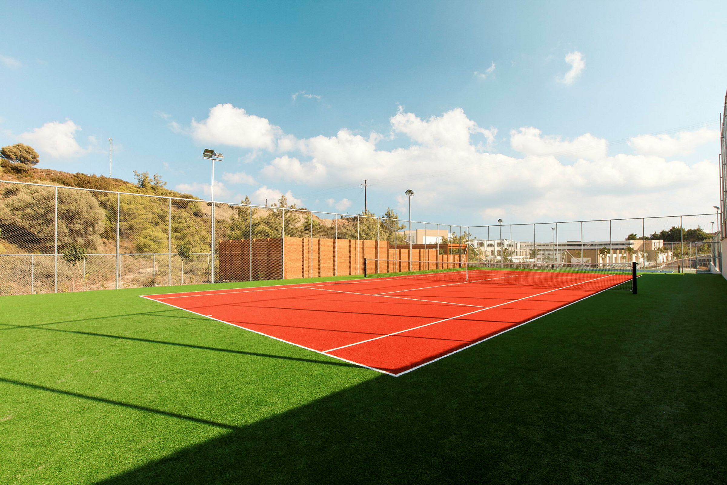 Kuva ulkotenniskentästä, jossa on tekonurmi ja verkko kirkkaan sinisen taivaan alla. (Tekoälyn luoma teksti)