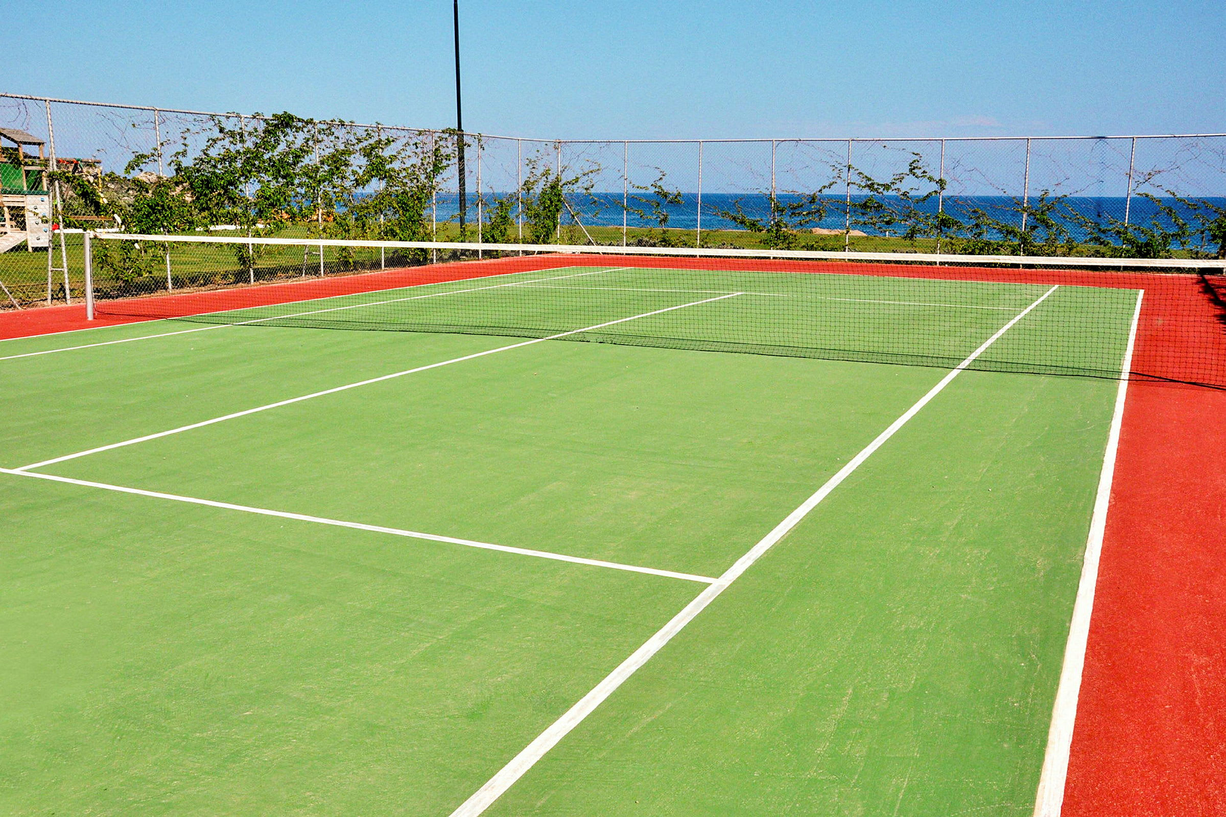 Tenniskenttä, jossa on vihreä pinta ja punaiset reunat, ympäröity verkkoaidalla ja kasvillisuudella, taustalla näkyy meri. (Tekoälyn luoma teksti)